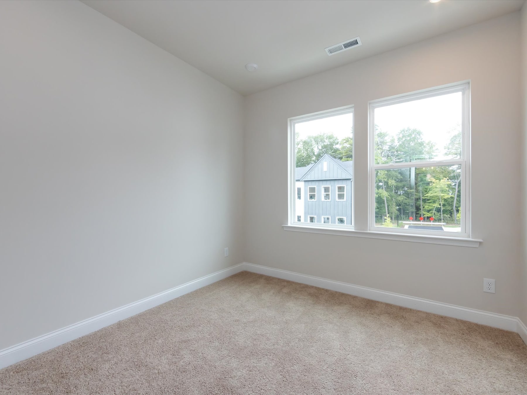 A bright, spacious room with large windows overlooking a residential neighborhood, featuring a neutral color scheme and plush carpeting.