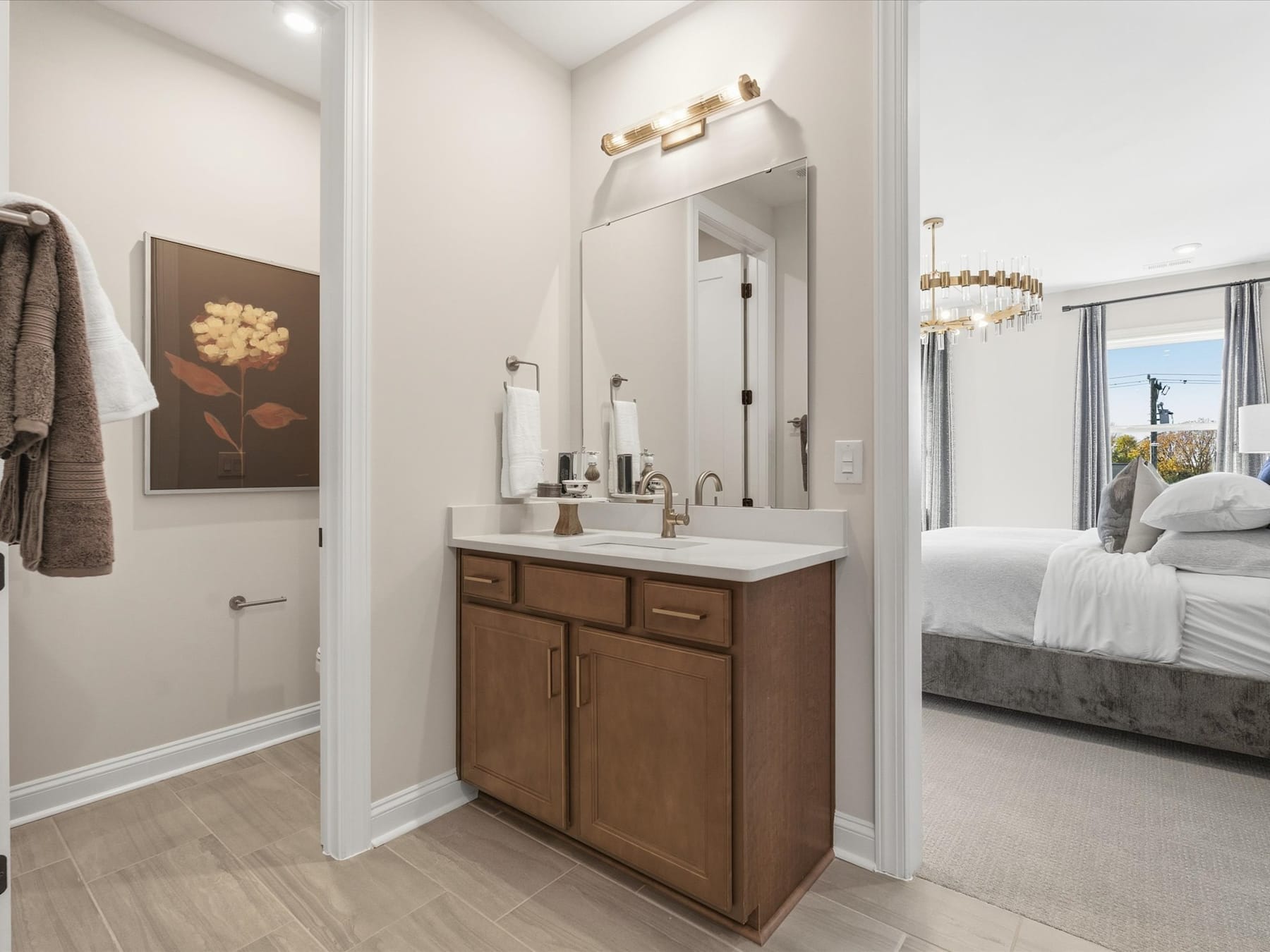 A modern bathroom with a wooden vanity, a large mirror, and a floral artwork on the wall, situated adjacent to a bedroom with a bed visible in the background.