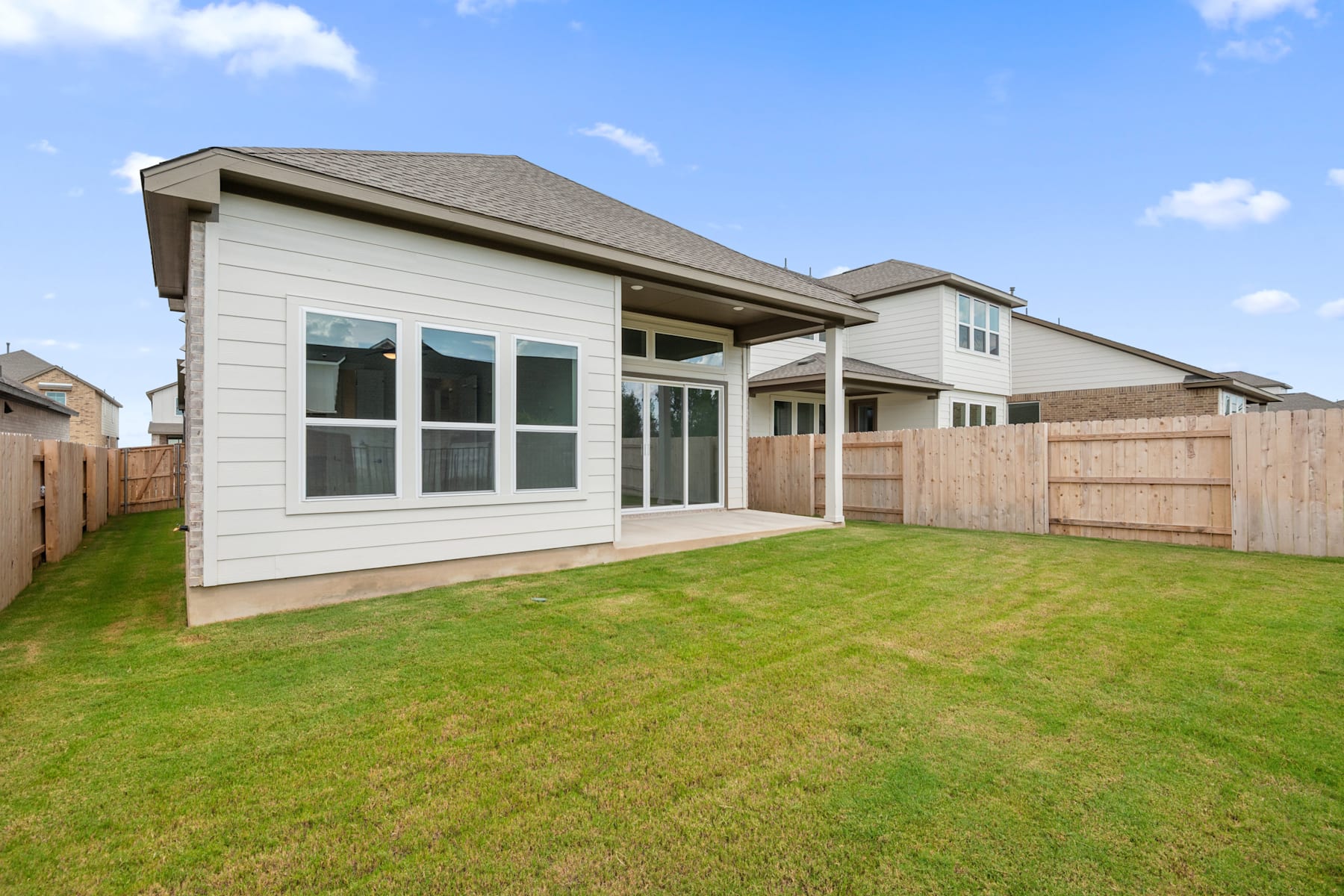 A modern two-story house with a covered patio stands in a well-manicured yard, surrounded by a wooden fence, against a backdrop of a blue sky with scattered clouds.
