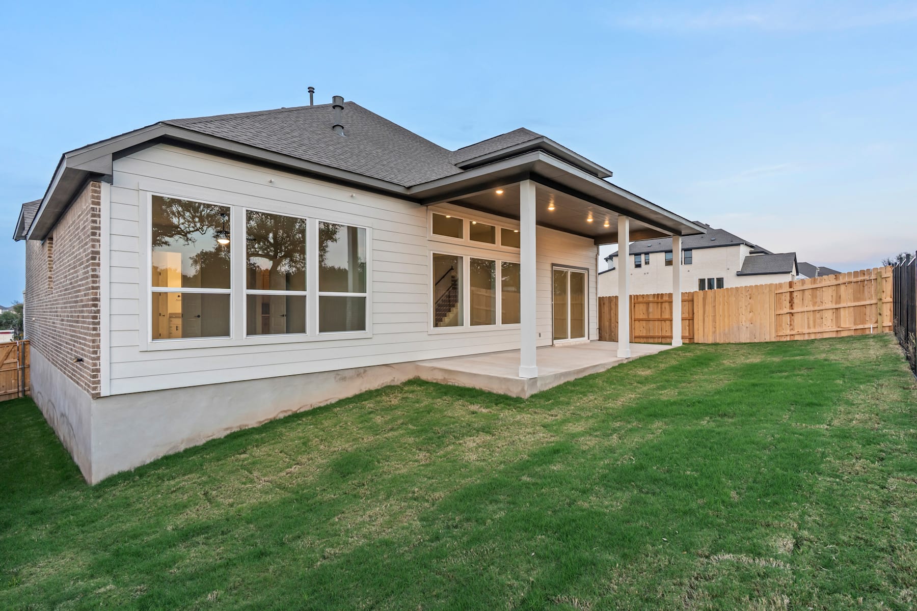 A modern two-story house with a large yard and a wooden fence in the foreground, set against a clear blue sky.