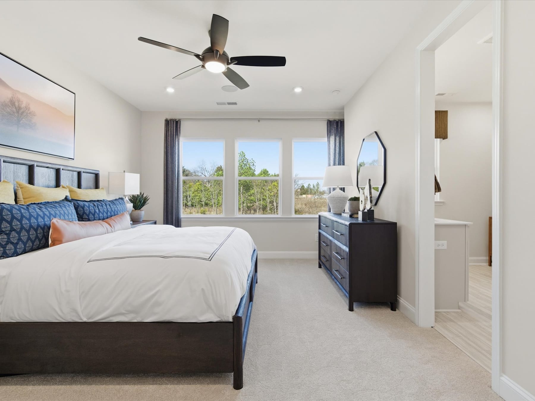 A spacious and well-lit bedroom with a large bed, dark furniture, and a ceiling fan, overlooking a scenic outdoor view through the windows.