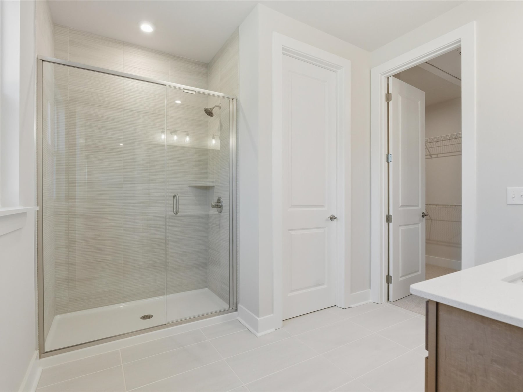 A modern, minimalist bathroom with a large glass shower enclosure, white walls, and a wooden vanity.