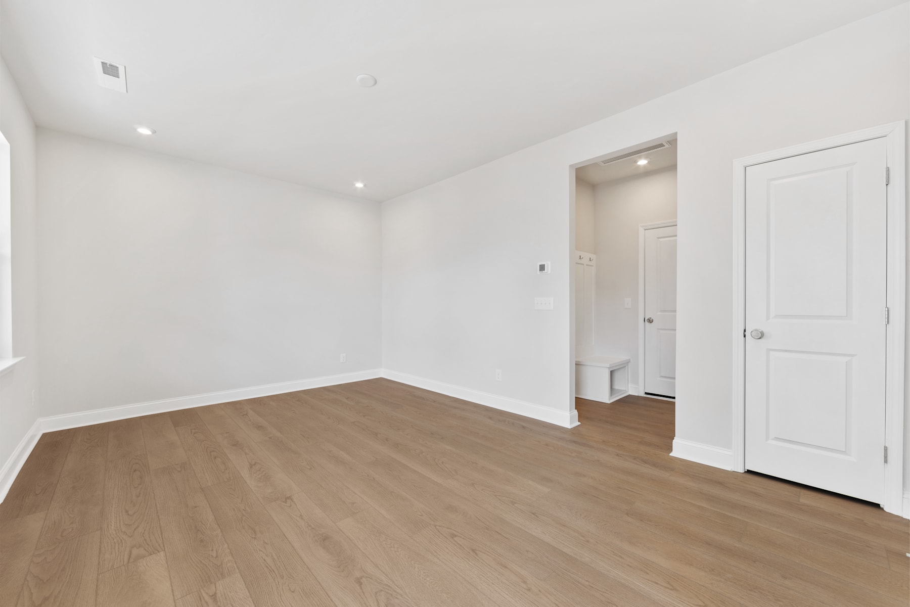 A spacious, well-lit room with white walls and a wooden floor, featuring a doorway leading to another room.