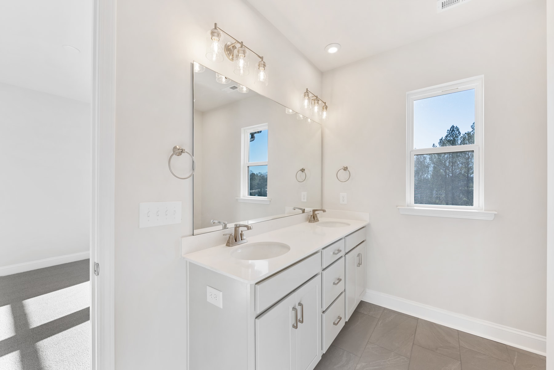 A modern, well-lit bathroom with a white vanity, double sinks, and a large mirror with wall-mounted lighting fixtures. The room features a window overlooking an outdoor scene.