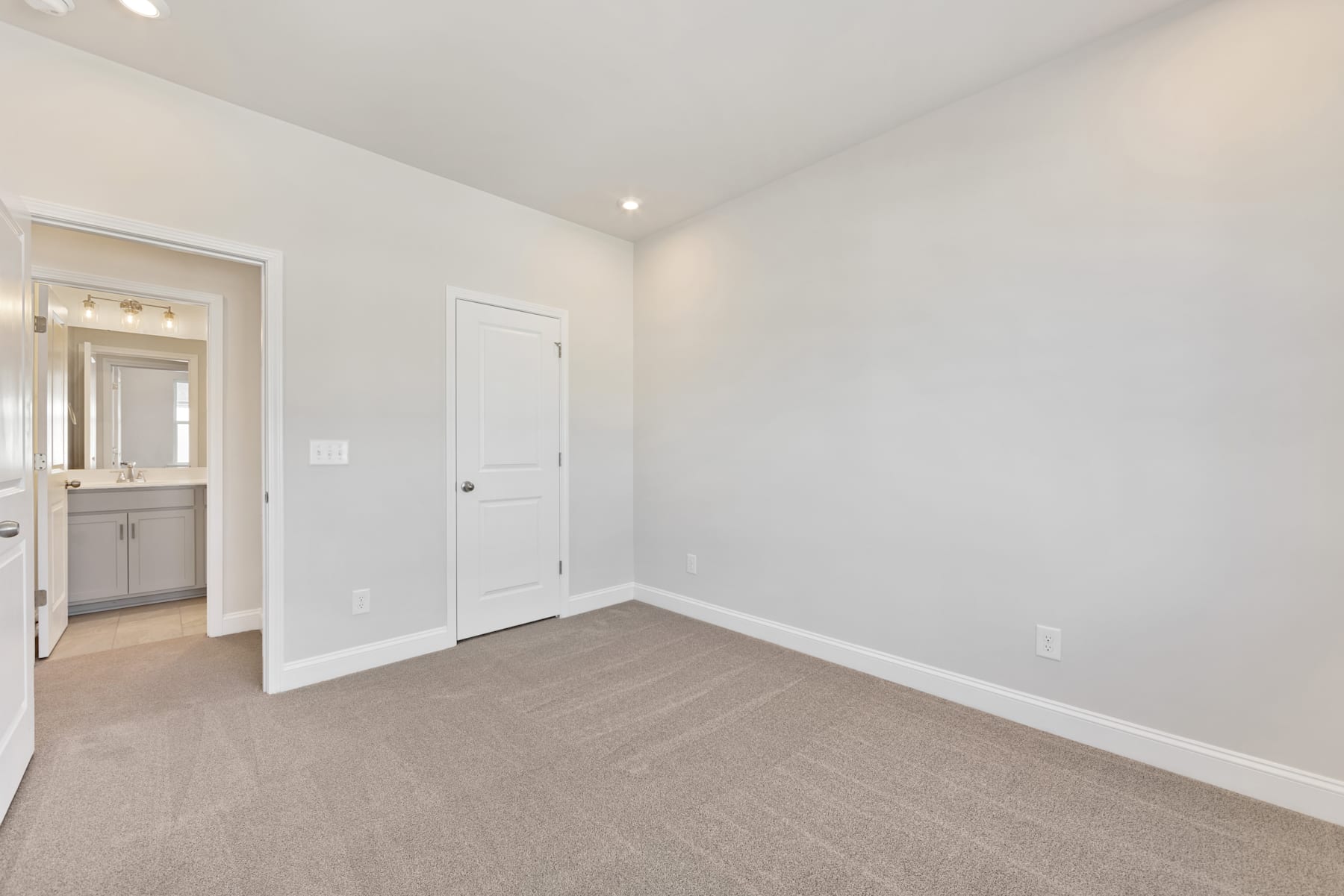 A spacious, well-lit room with white walls, a carpeted floor, and a partially visible bathroom door in the background.