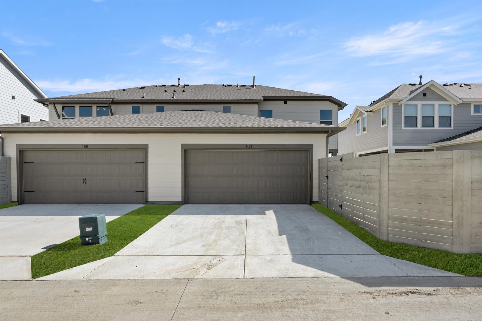 A modern two-story house with a garage, surrounded by a grassy yard and a concrete driveway, set against a clear blue sky with some clouds.