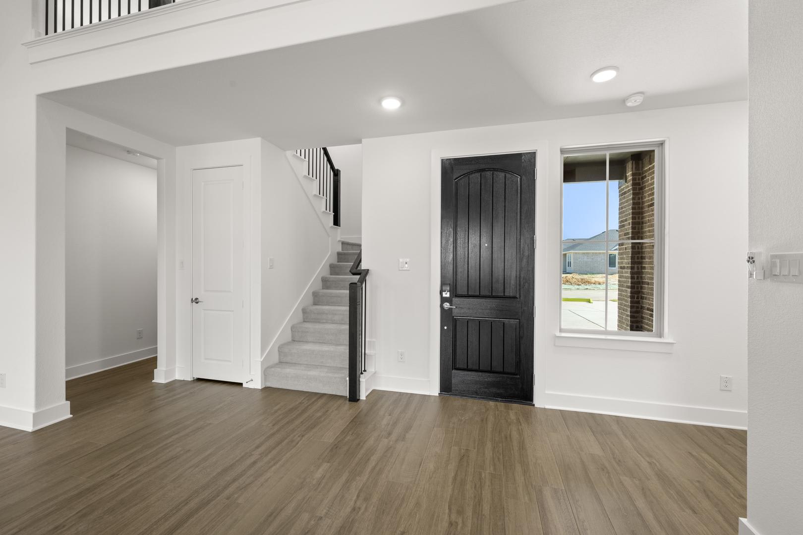 A spacious and well-lit interior with a dark wooden door, hardwood floors, and a staircase leading to the upper level.