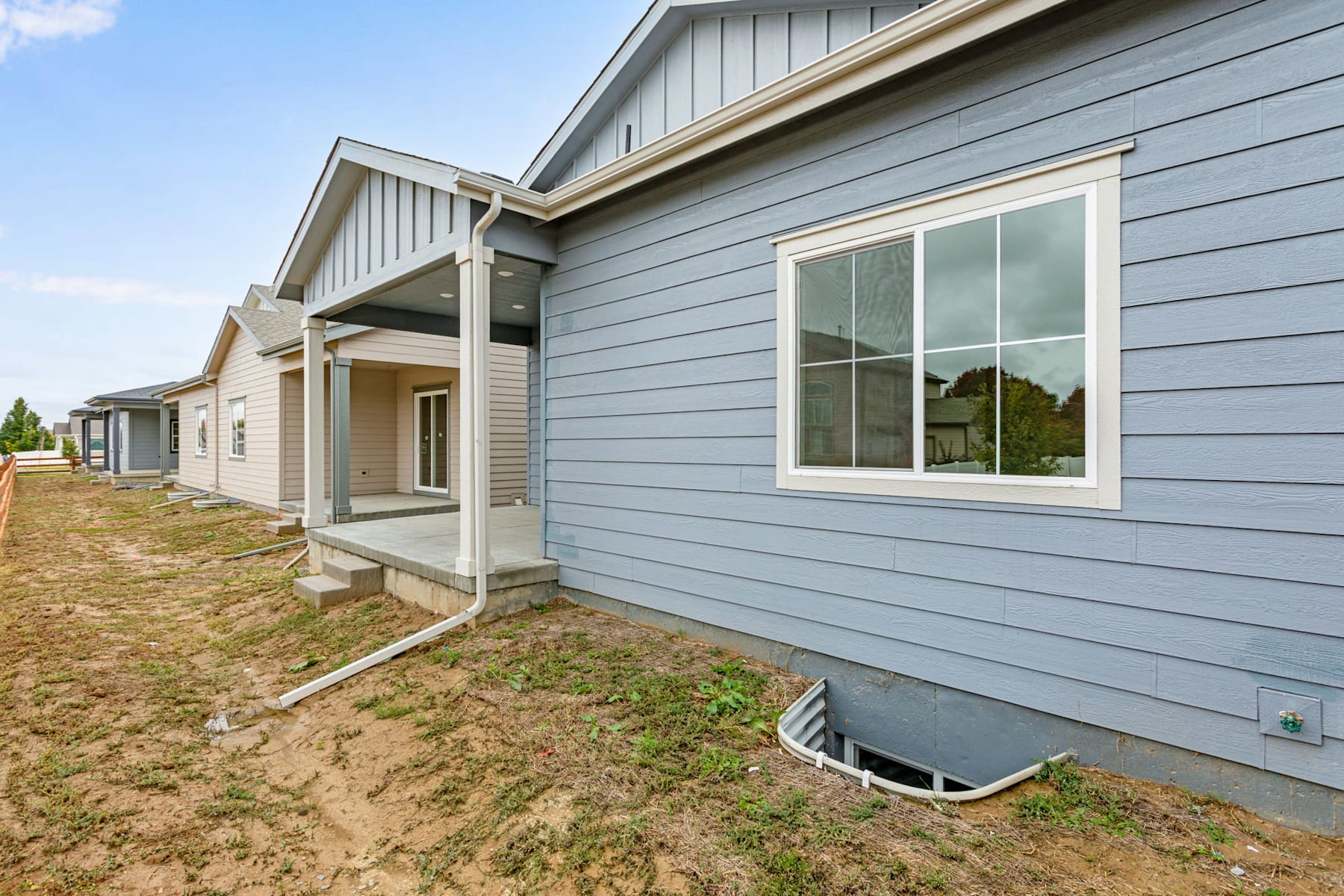 The image shows a row of newly constructed houses with gray siding, large windows, and covered porches. The foreground features a grassy area with a small foundation or basement visible.