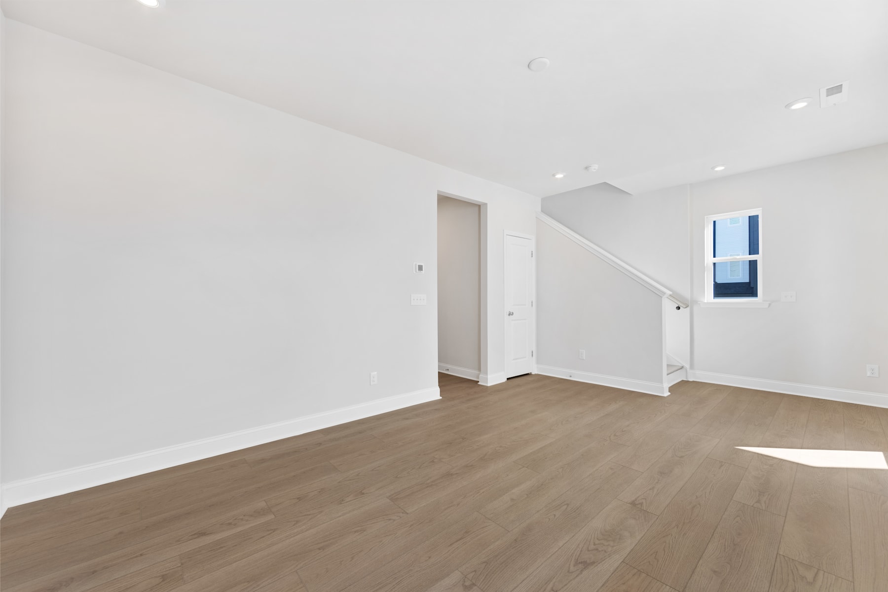 A spacious, minimalist room with white walls, wooden flooring, and a window providing natural light.