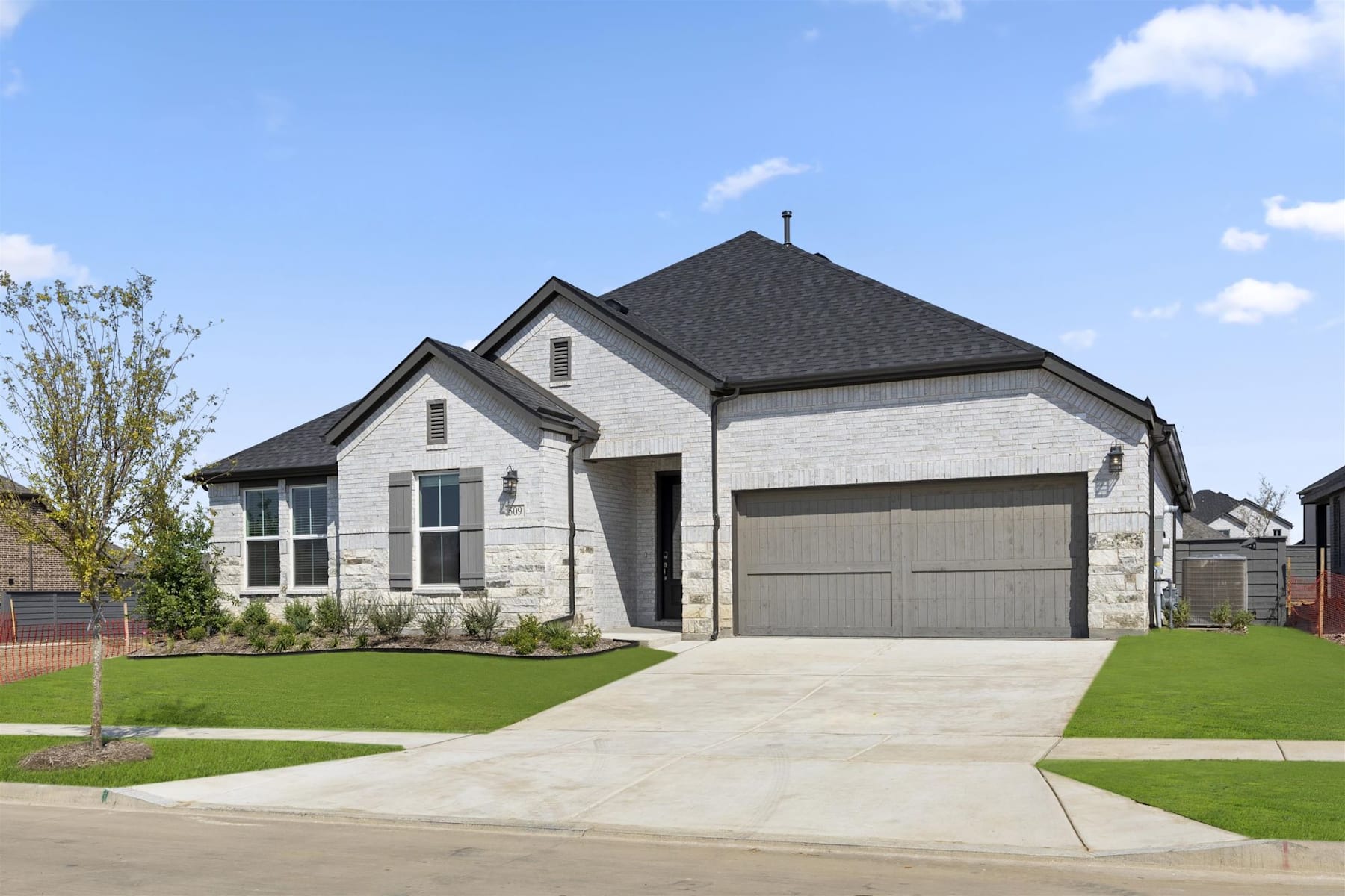 A modern two-story house with a gray roof, white siding, and a garage door sits on a well-manicured lawn surrounded by lush greenery and a clear blue sky with fluffy white clouds.