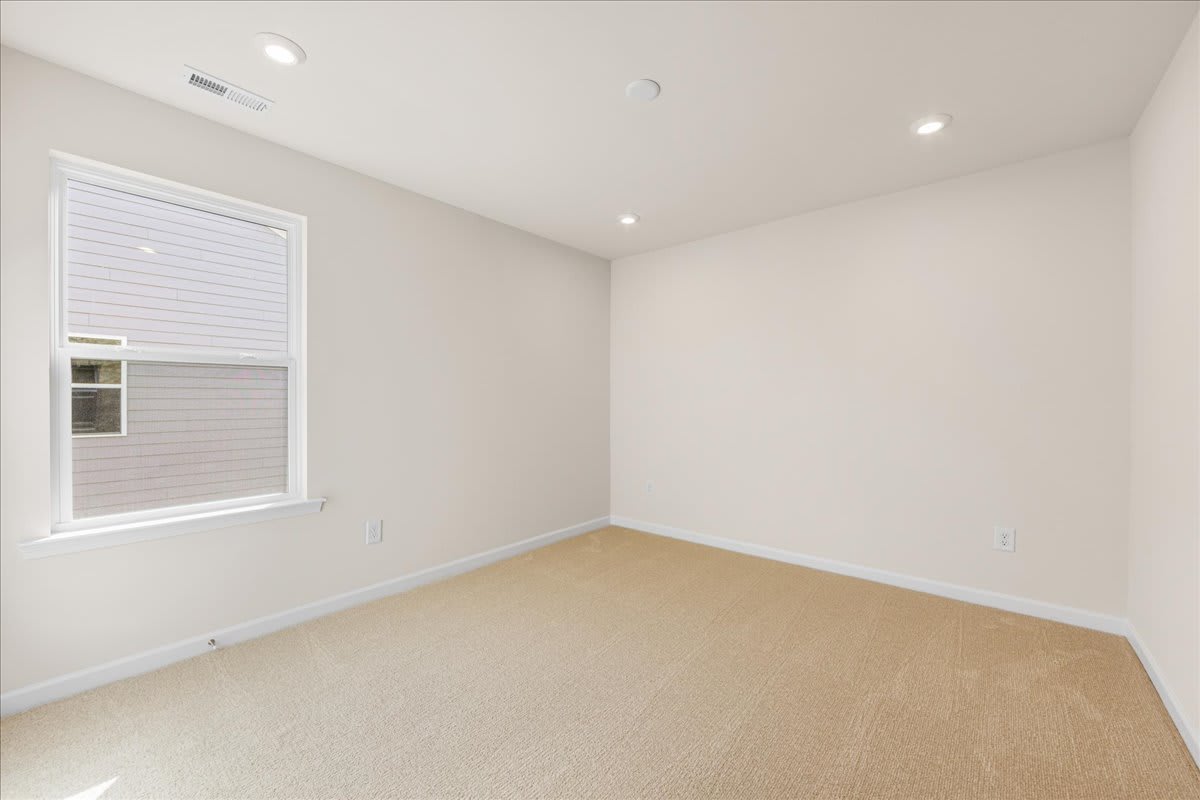 A bright, empty room with white walls, a window, and a beige carpeted floor.