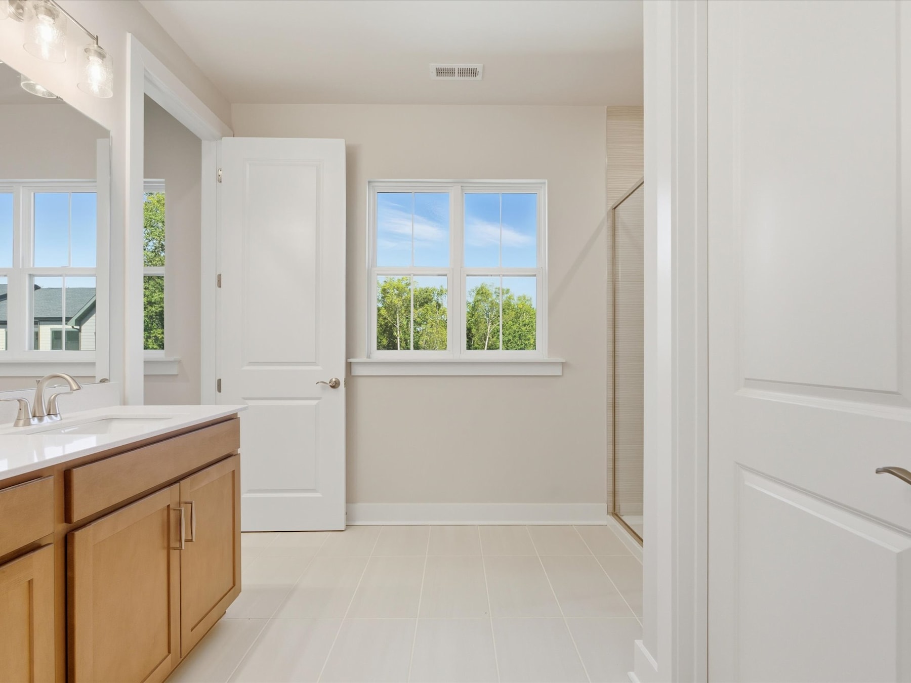 A bright and airy bathroom with a wooden vanity, white walls, and large windows overlooking a lush, green outdoor scene.
