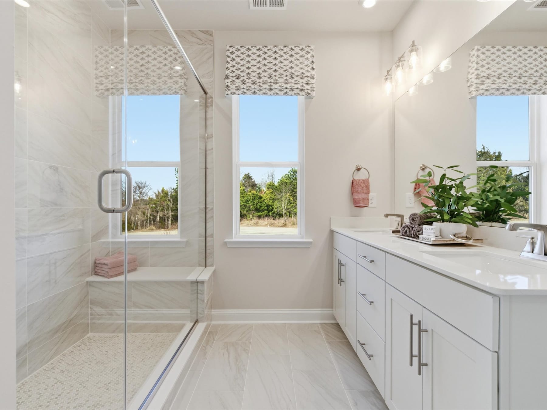 A bright and spacious bathroom with a glass-enclosed shower, white cabinets, and large windows overlooking a scenic outdoor landscape.