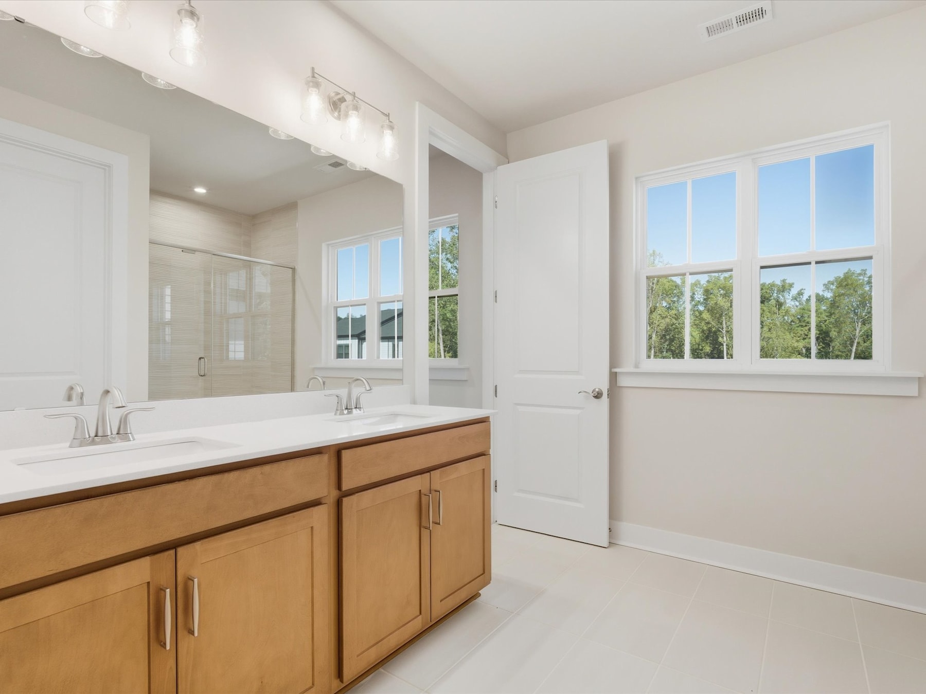 A spacious and well-lit bathroom with a wooden vanity, white countertops, and large windows overlooking a lush outdoor landscape.