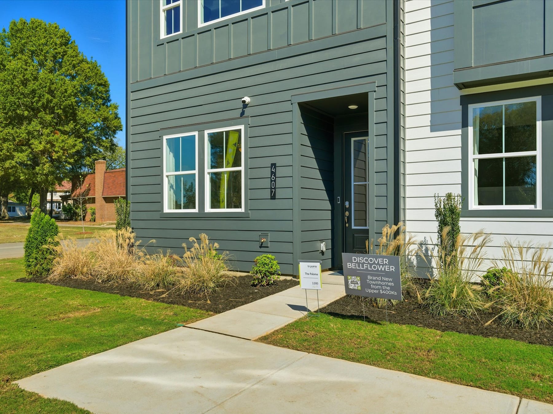 The image shows a modern, multi-story building with a gray exterior and large windows. The foreground features a paved walkway leading to the building's entrance, surrounded by landscaping with small shrubs and plants.