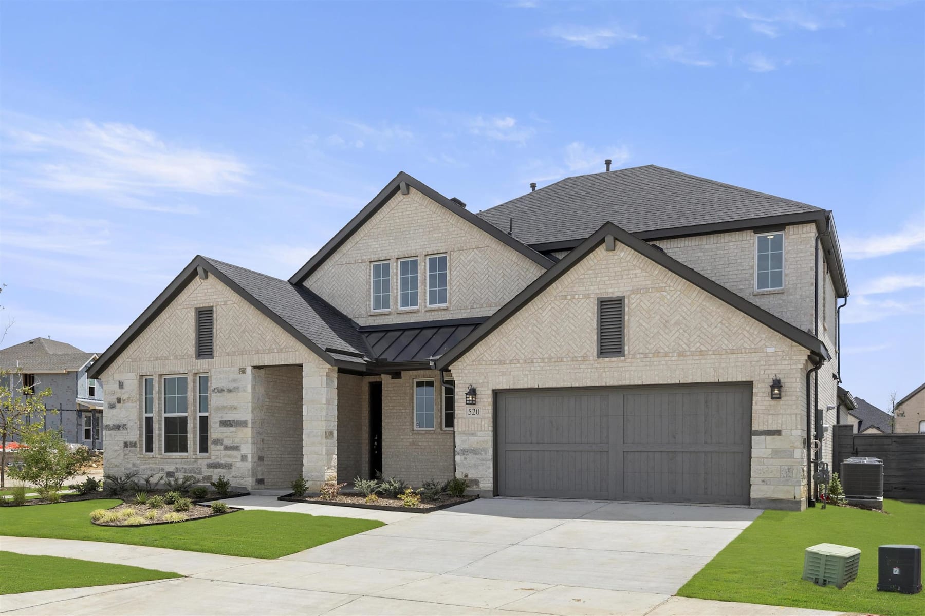 A two-story residential house with a gray roof, beige exterior, and a well-manicured lawn in the foreground, set against a clear blue sky with scattered clouds.