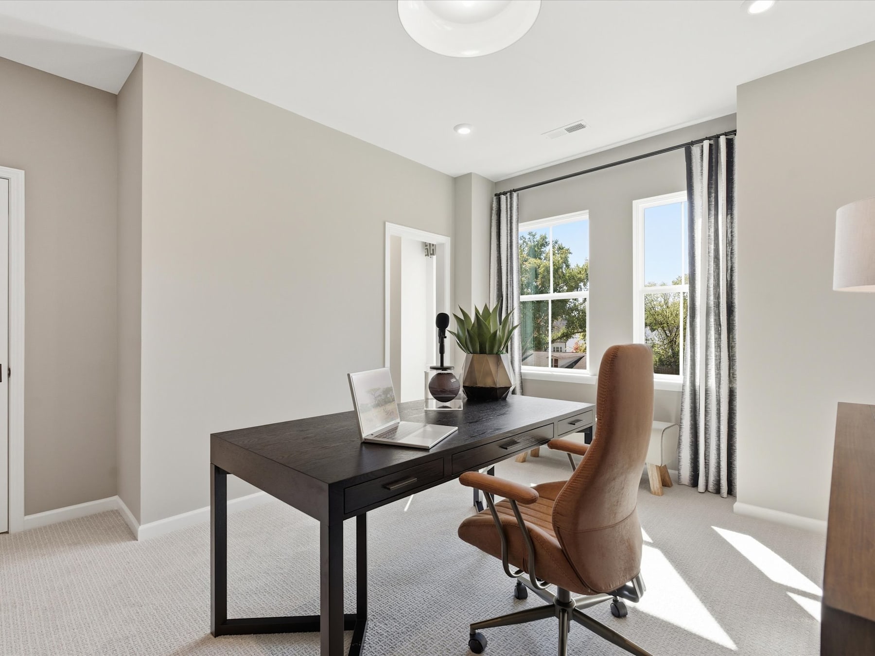 A modern and minimalist home office space with a dark wooden desk, a comfortable upholstered chair, and large windows providing natural light and a view of the outdoor landscape.