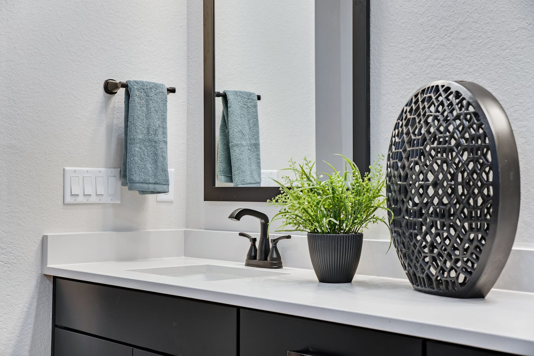 A modern bathroom vanity with a black cabinet, white countertop, and decorative vase with greenery, framed by mirrors and towel racks on the wall.