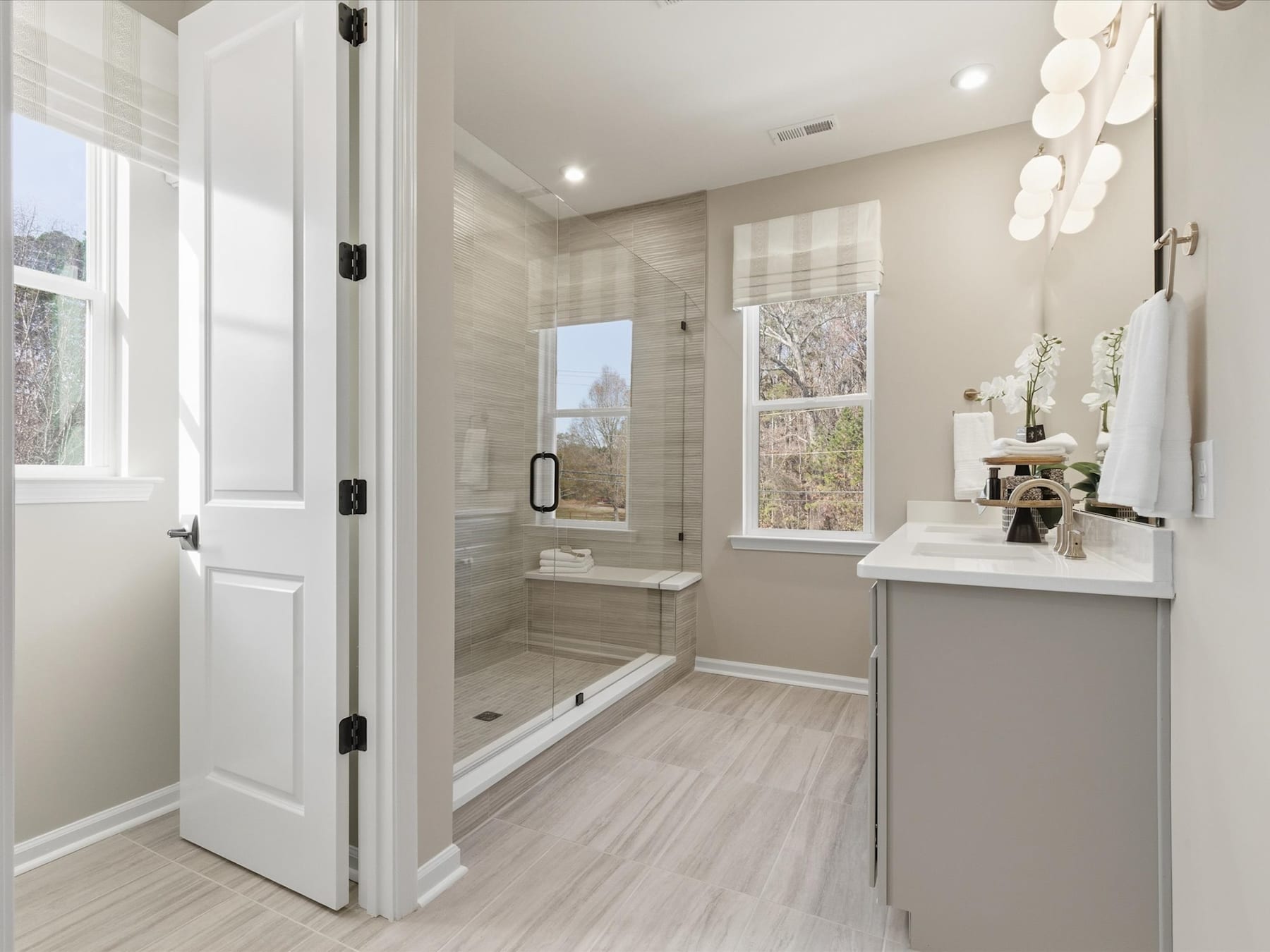 A spacious and well-lit bathroom with a modern design, featuring a large window, a vanity with a white countertop, and a glass-enclosed shower area.