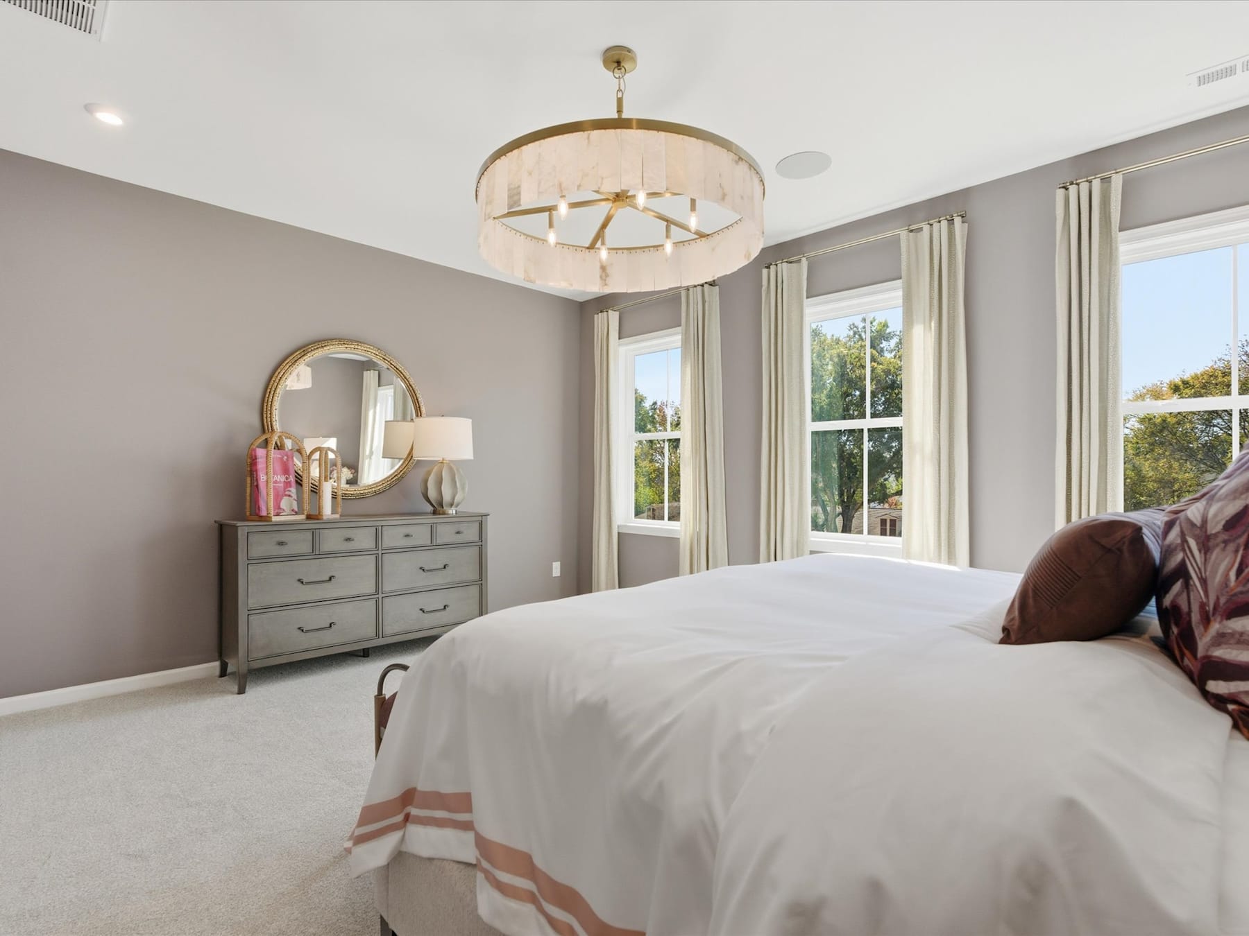 A spacious and elegantly decorated bedroom with a large bed, a dresser, and a chandelier hanging from the ceiling, surrounded by large windows that provide natural light.