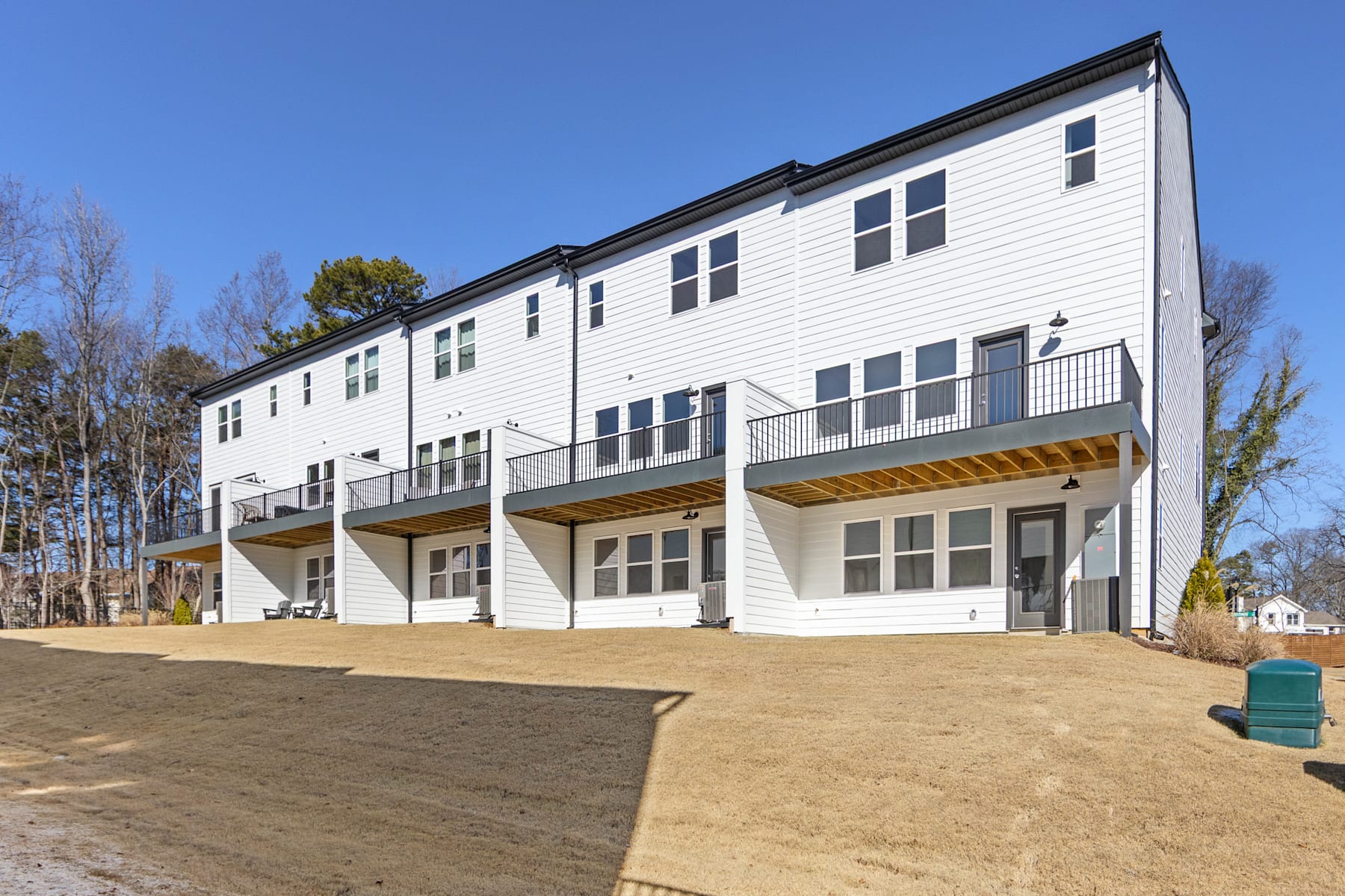 A row of modern, white townhouses with balconies and a grassy area in the foreground, set against a backdrop of trees and a clear blue sky.