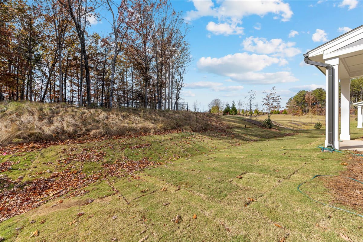 A grassy slope leads up to a wooded area with tall trees under a cloudy blue sky, with a white building visible in the background.