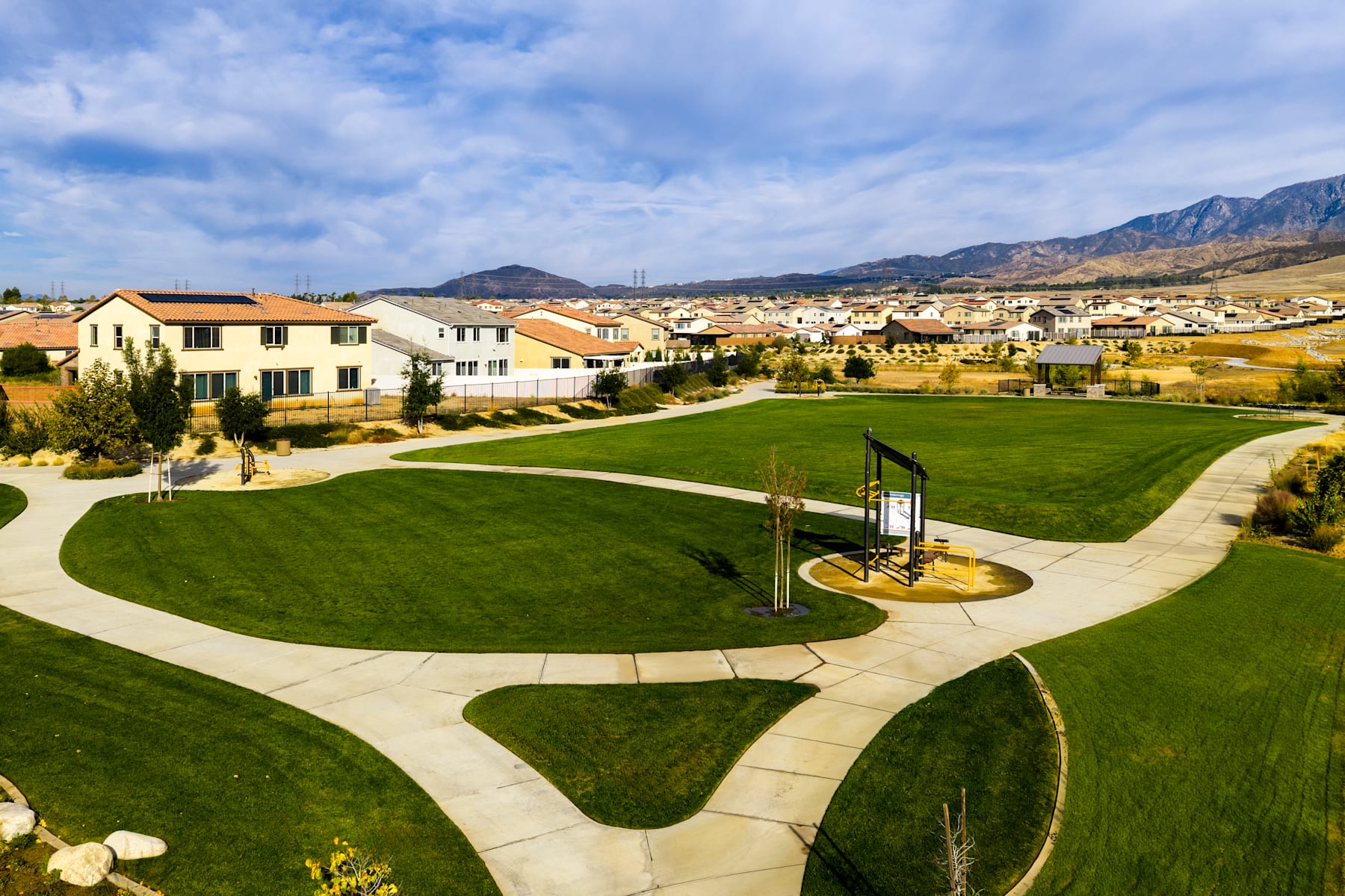 A well-manicured lawn with winding paths and a gazebo-like structure in the foreground, surrounded by a residential neighborhood nestled against a mountainous backdrop under a cloudy sky.
