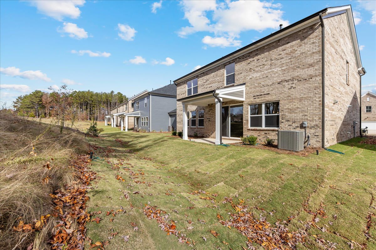 The image shows a row of modern brick townhouses with a grassy lawn in the foreground and a wooded area in the background.