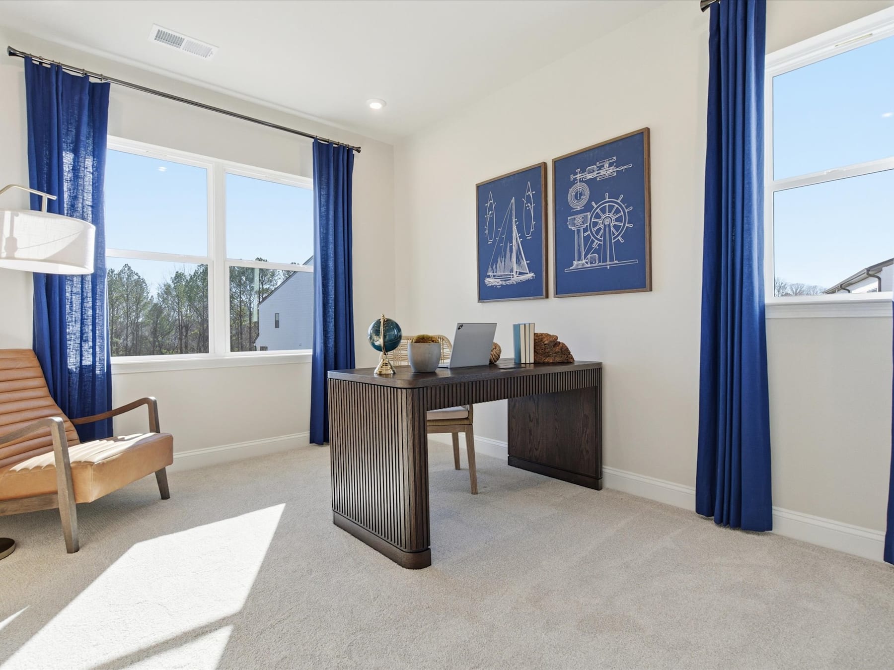 A modern and minimalist home office space with a wooden desk, navy blue curtains, and framed artwork on the walls, overlooking a scenic outdoor view through the large windows.