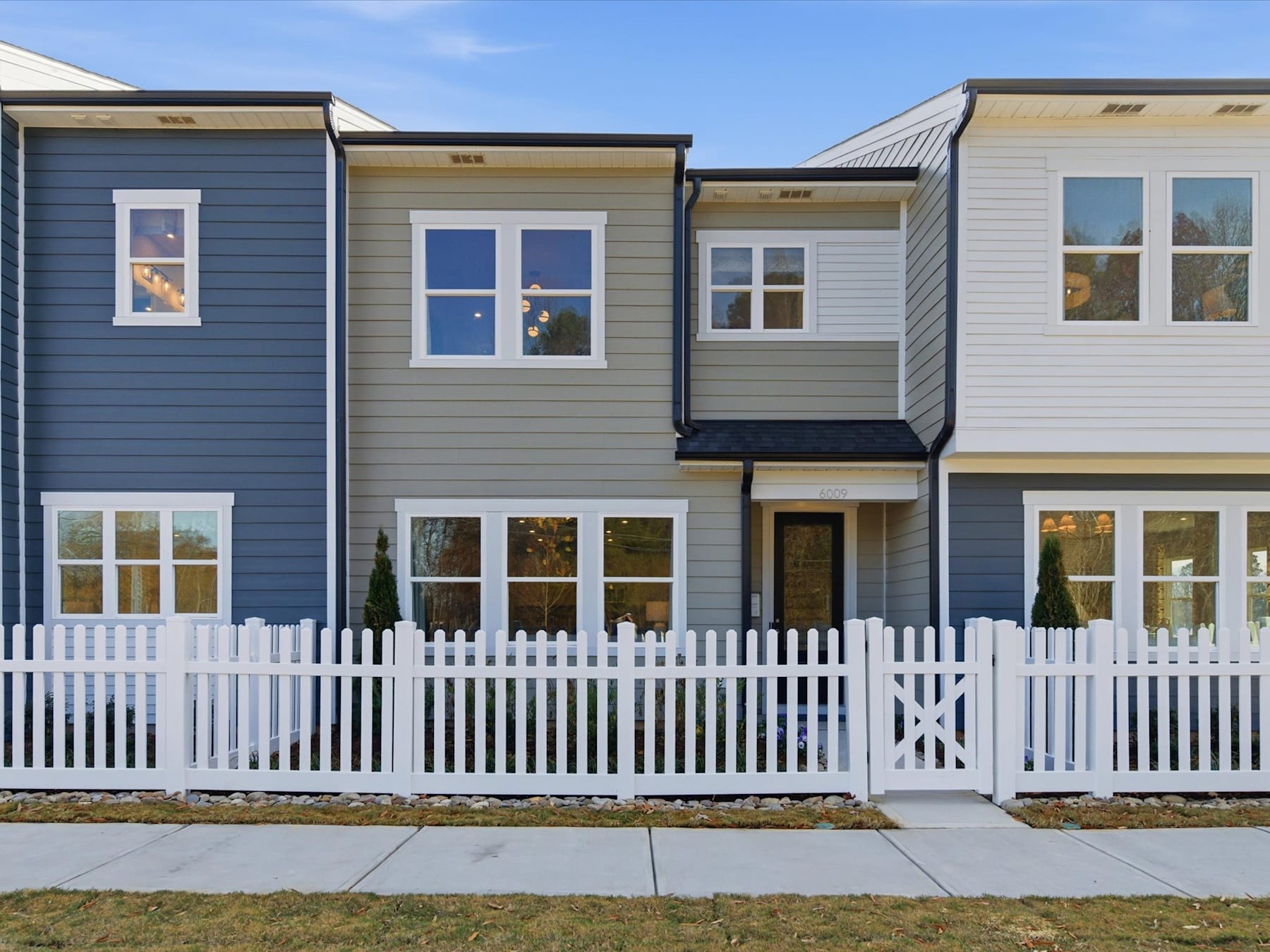 A row of colorful, modern townhouses with white picket fences in the foreground, set against a clear blue sky in the background.