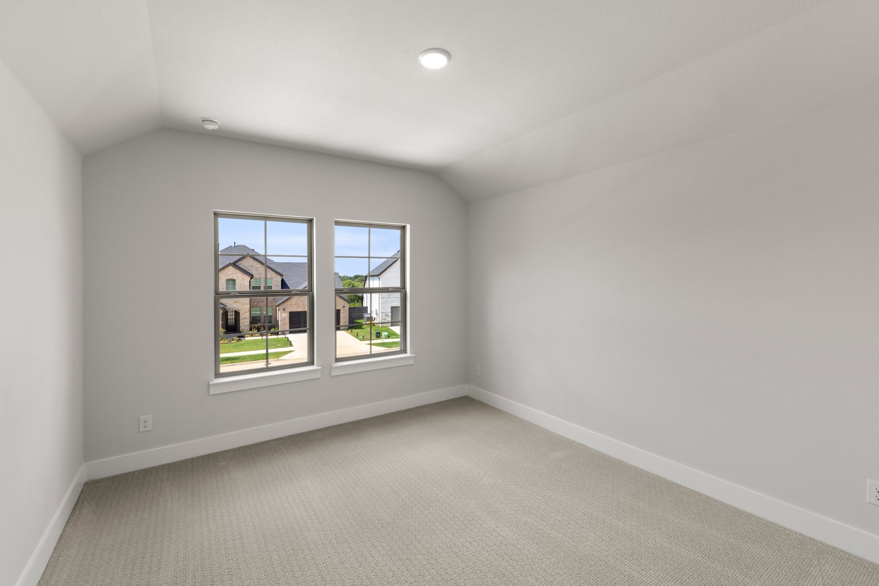 An empty room with large windows overlooking a residential neighborhood, featuring a neutral color scheme and carpeted flooring.