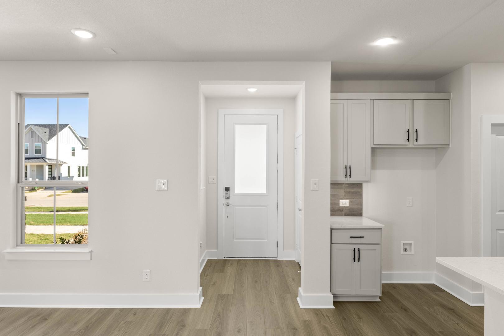 A bright and spacious interior with a white door, cabinets, and hardwood flooring, overlooking a grassy outdoor area through a large window.