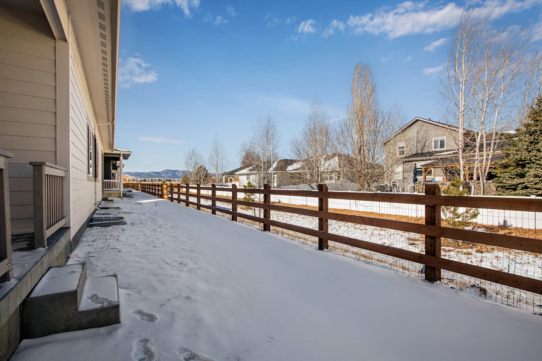 A snowy residential neighborhood with a wooden fence in the foreground and houses, trees, and a clear blue sky in the background.