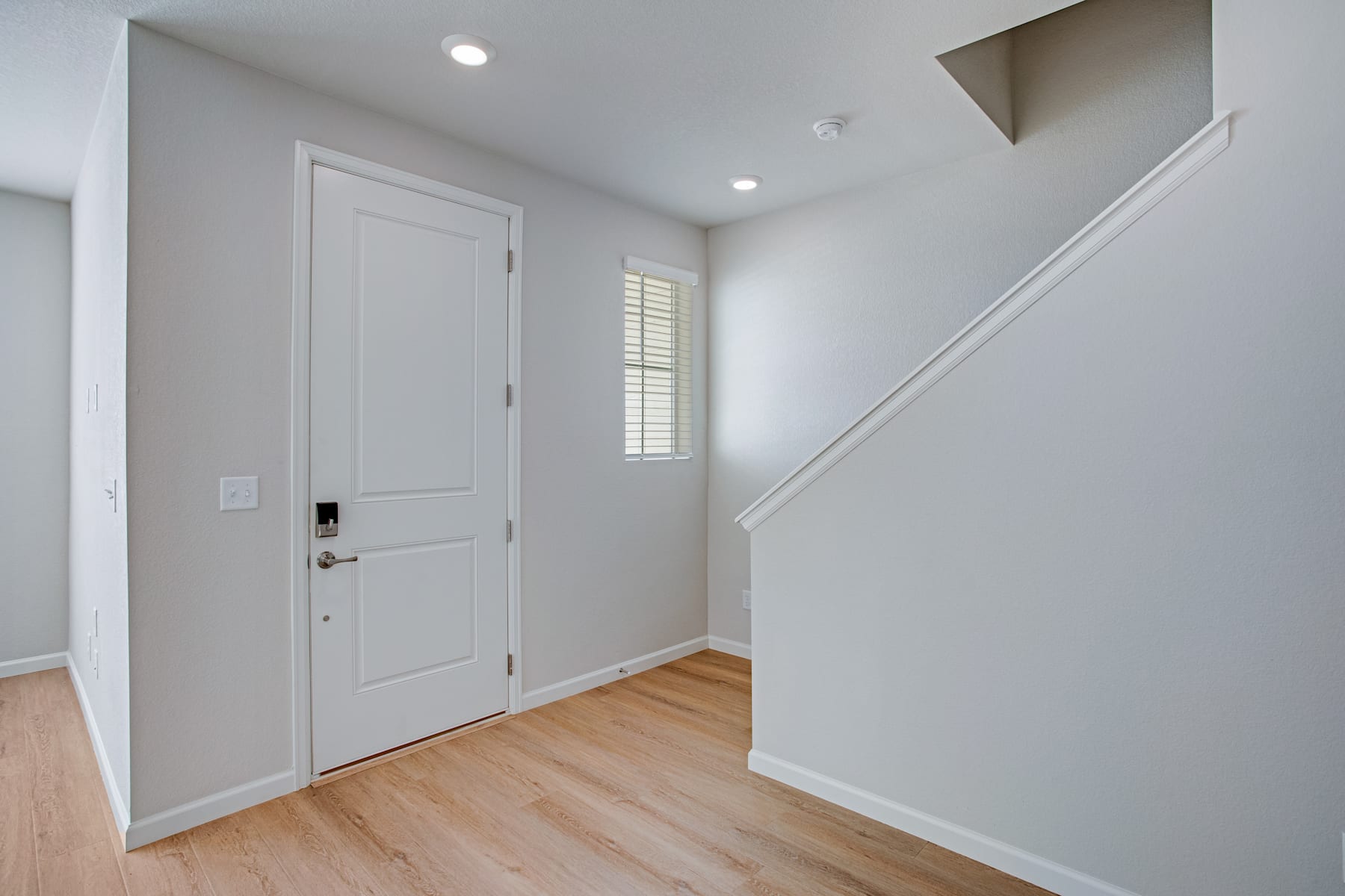 A bright and airy entryway with a white door, hardwood floors, and recessed lighting, leading to a staircase in the background.
