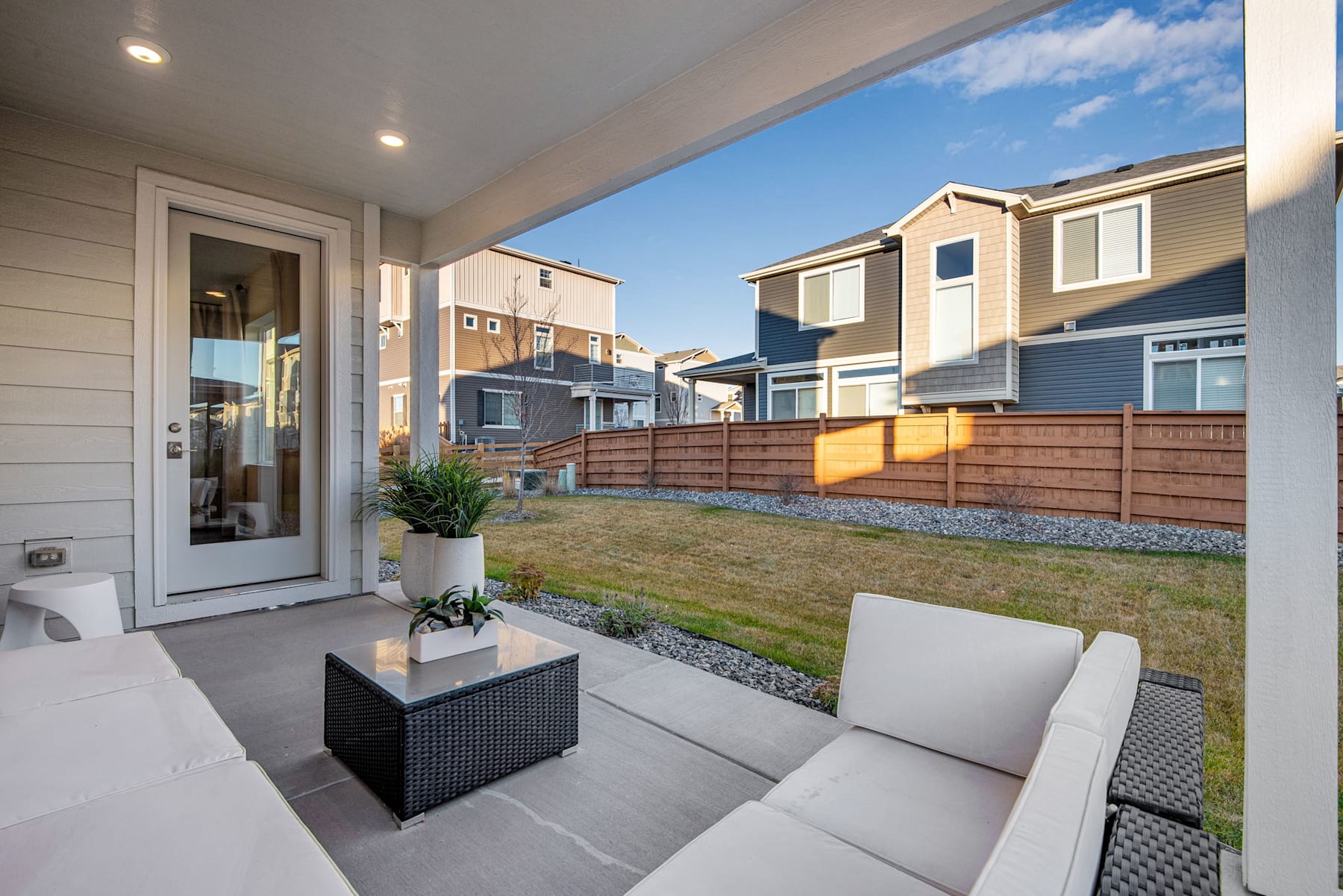 A cozy outdoor patio area with a wicker furniture set, surrounded by a well-maintained lawn and neighboring townhouses with a clear blue sky in the background.