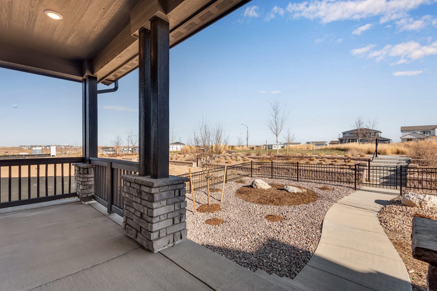 A covered porch with a brick column leads to a landscaped yard with gravel, rocks, and bare trees in the background, set against a clear blue sky.