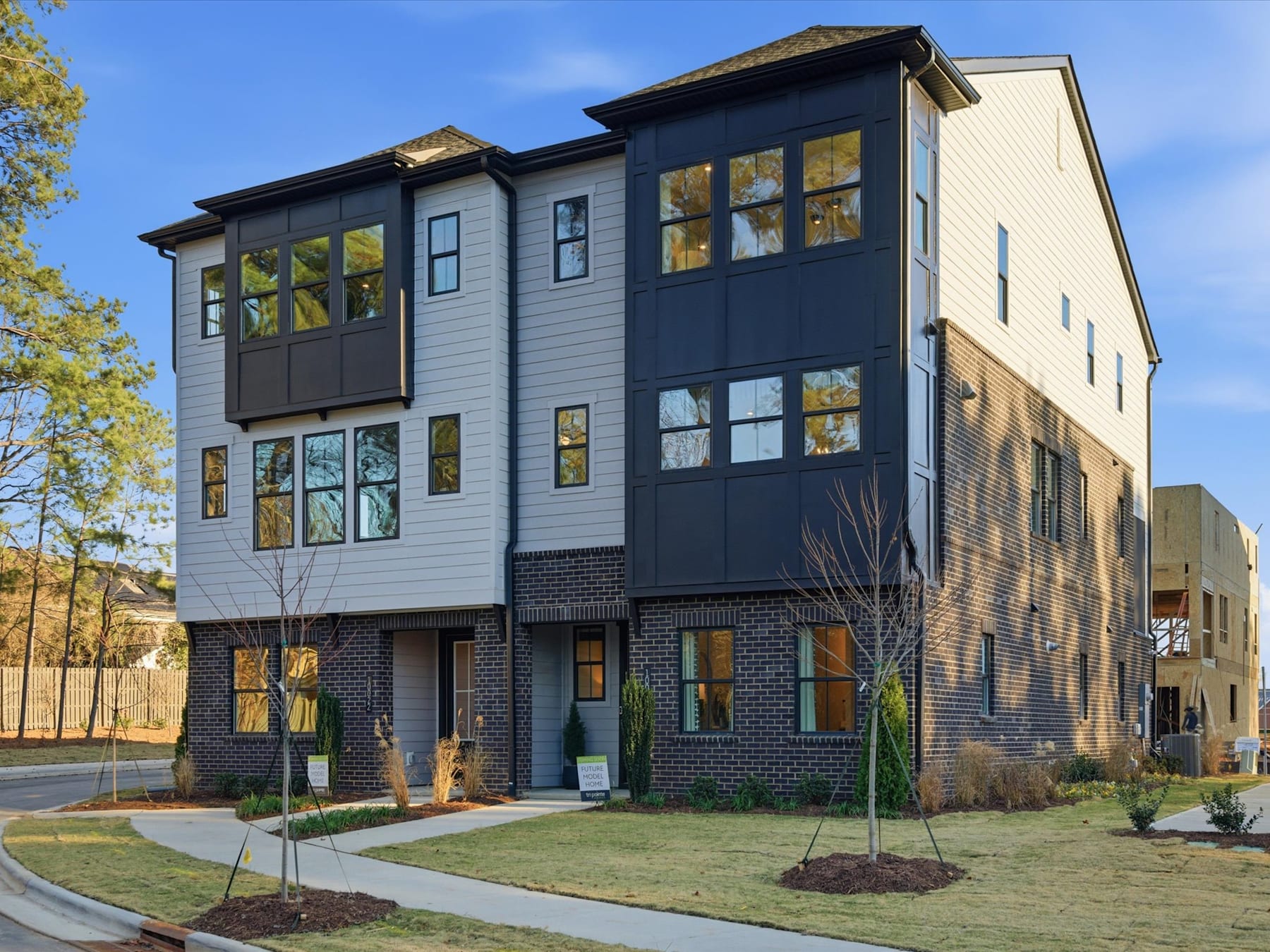 A modern, multi-story residential building with a mix of dark and light siding, surrounded by a well-landscaped yard with trees and a paved walkway in the foreground.