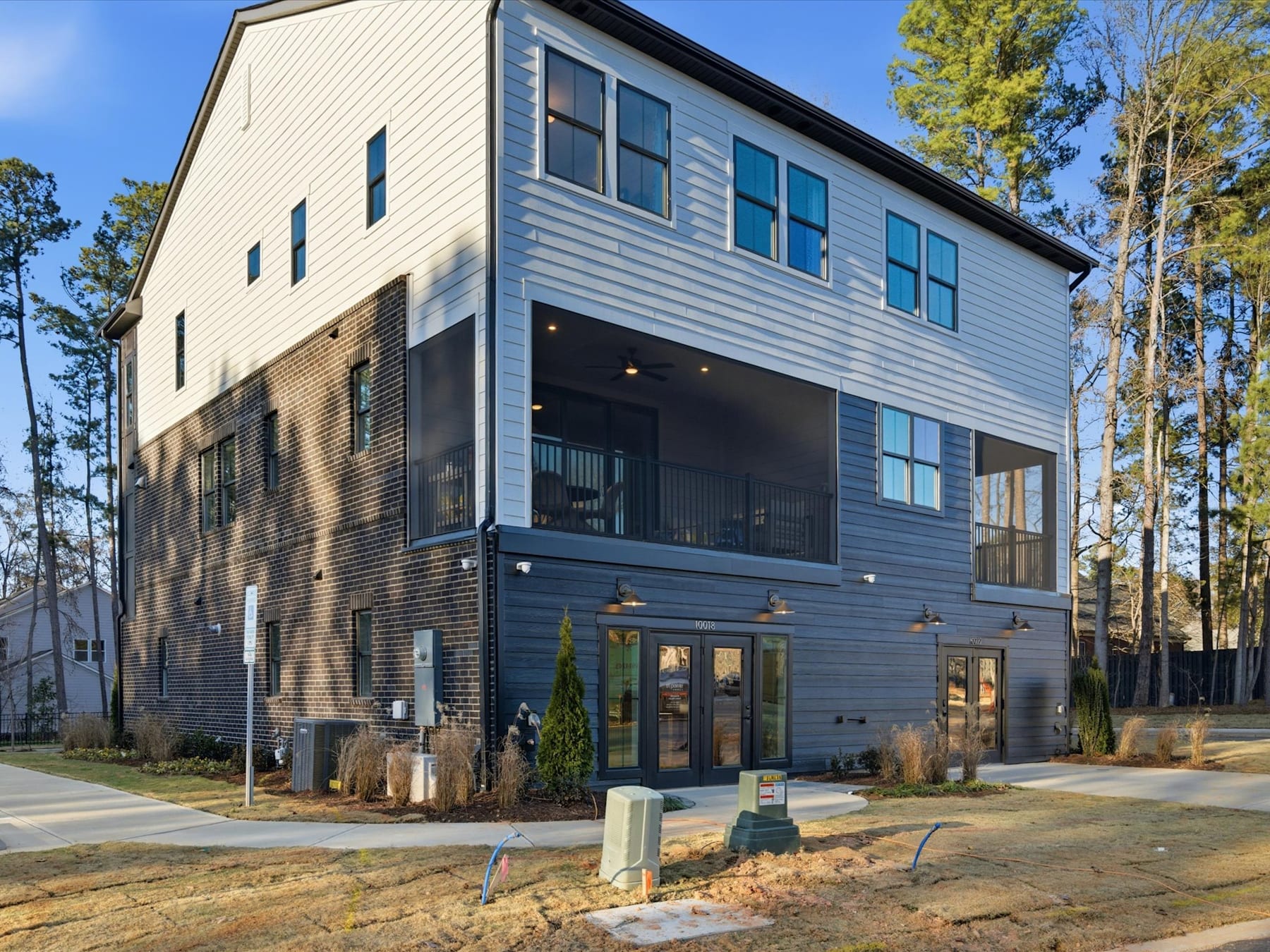 A modern, multi-story residential building with a mix of siding materials stands in a wooded area, with a paved walkway and landscaping in the foreground.