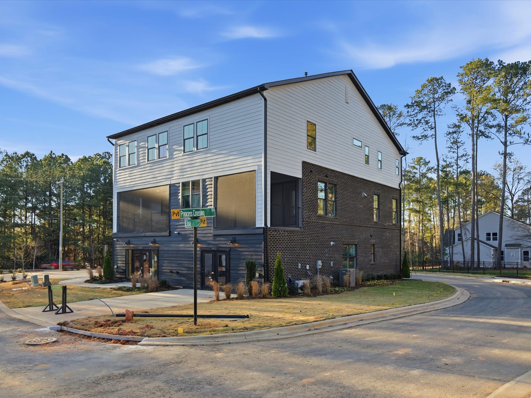 A modern two-story residential building with a mix of siding materials stands in a wooded area, surrounded by a paved road and landscaping.