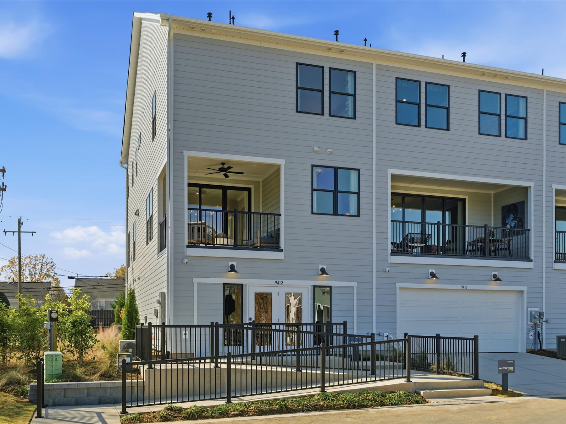 A multi-story residential building with a gray exterior, balconies, and a fenced-in yard in the foreground, set against a clear blue sky.