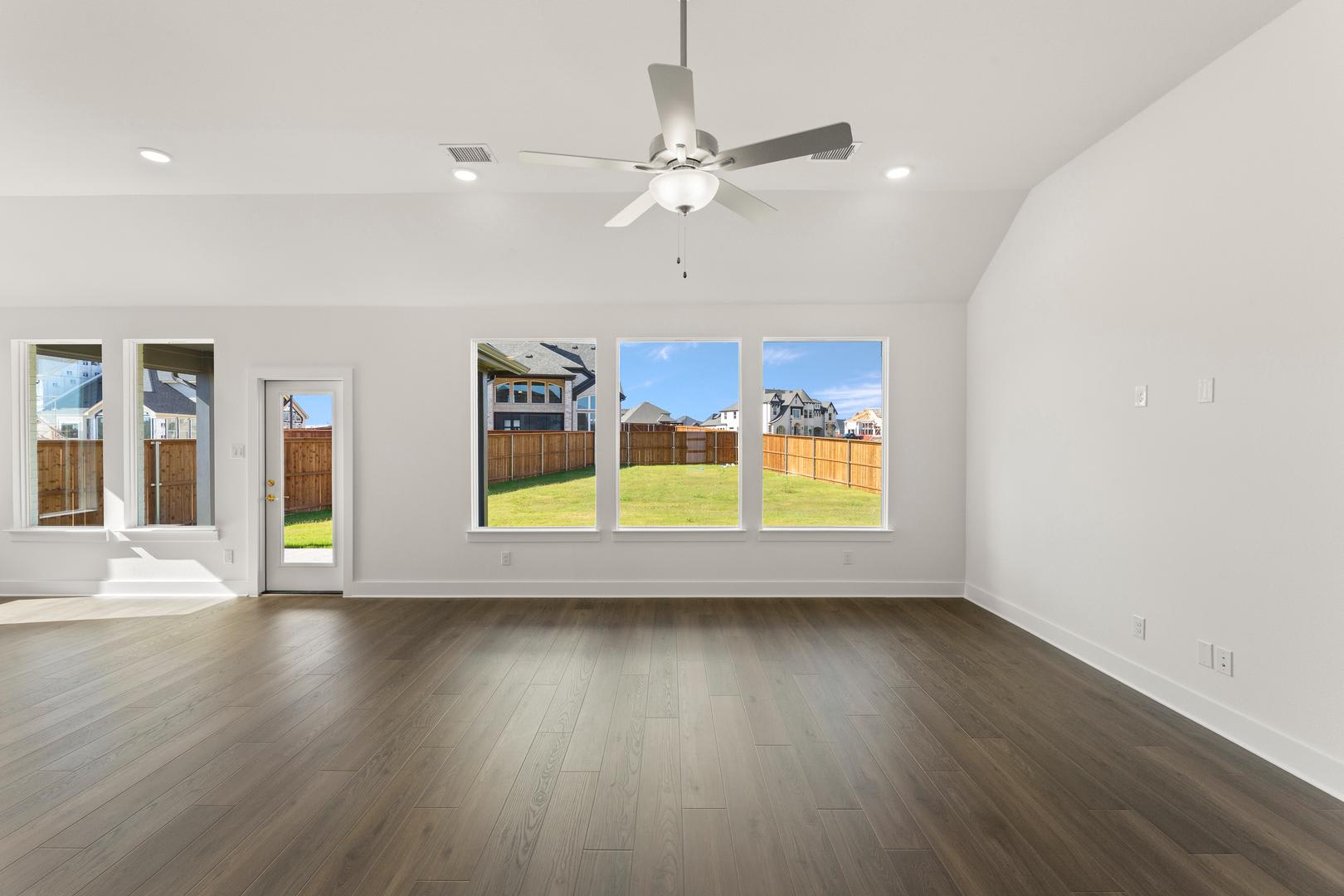 A spacious, well-lit room with a ceiling fan, hardwood floors, and large windows overlooking a backyard with a wooden fence.