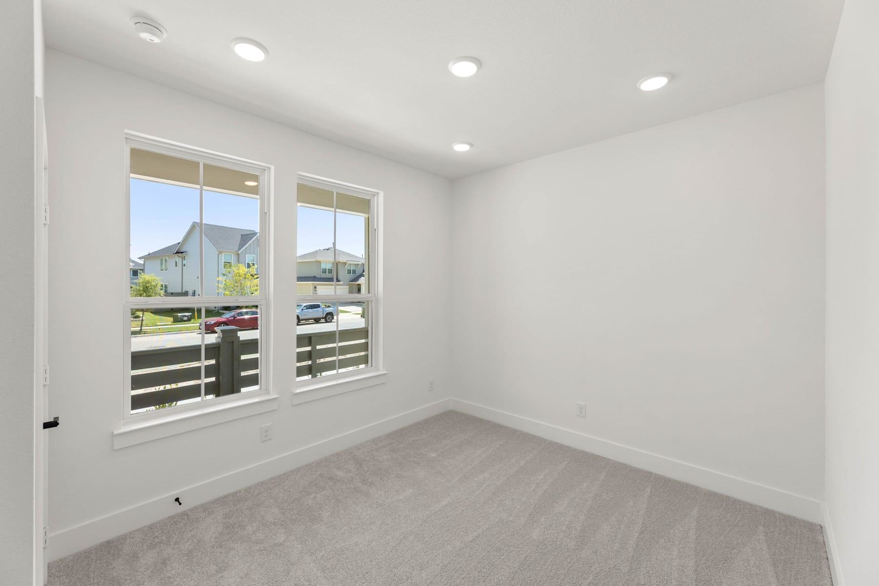 A bright, empty room with white walls, recessed lighting, and large windows overlooking a residential neighborhood in the background.