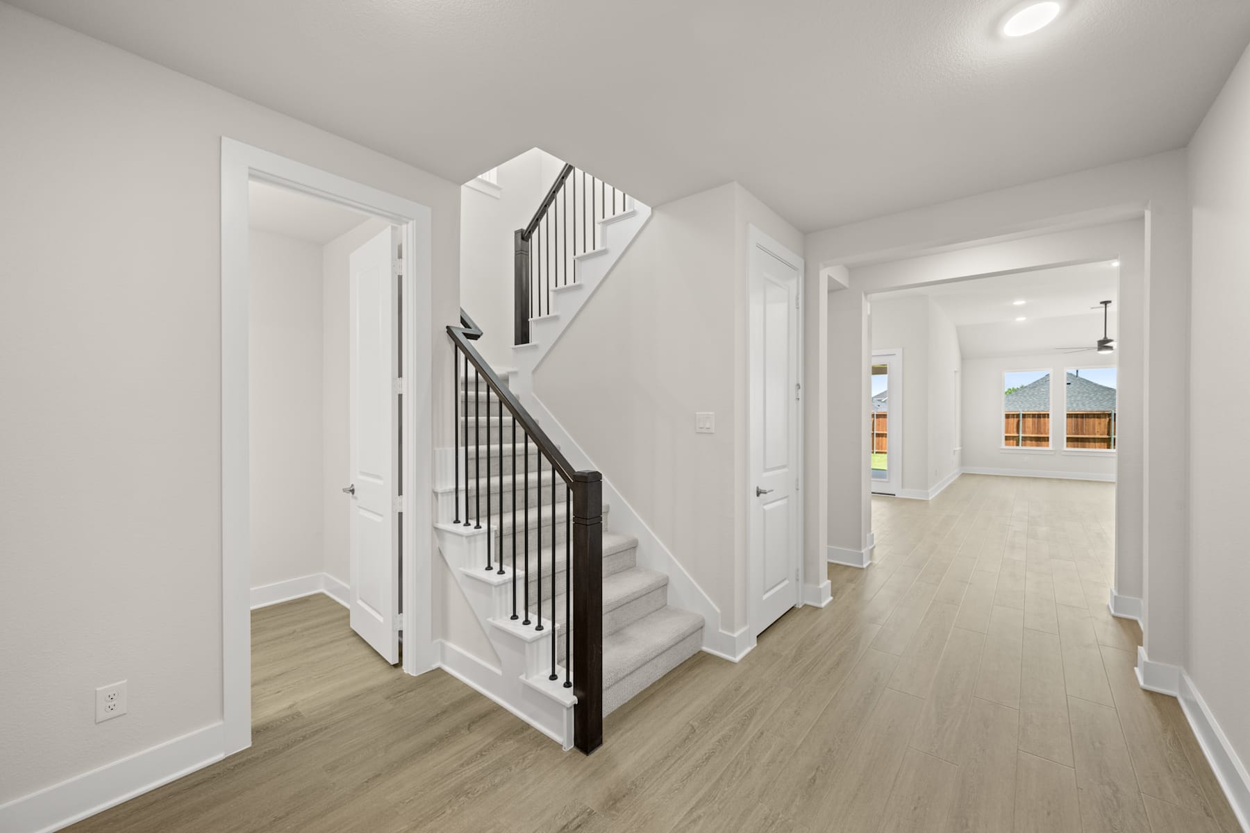 A bright, open hallway with white walls, a wooden staircase, and a view of a room in the background.