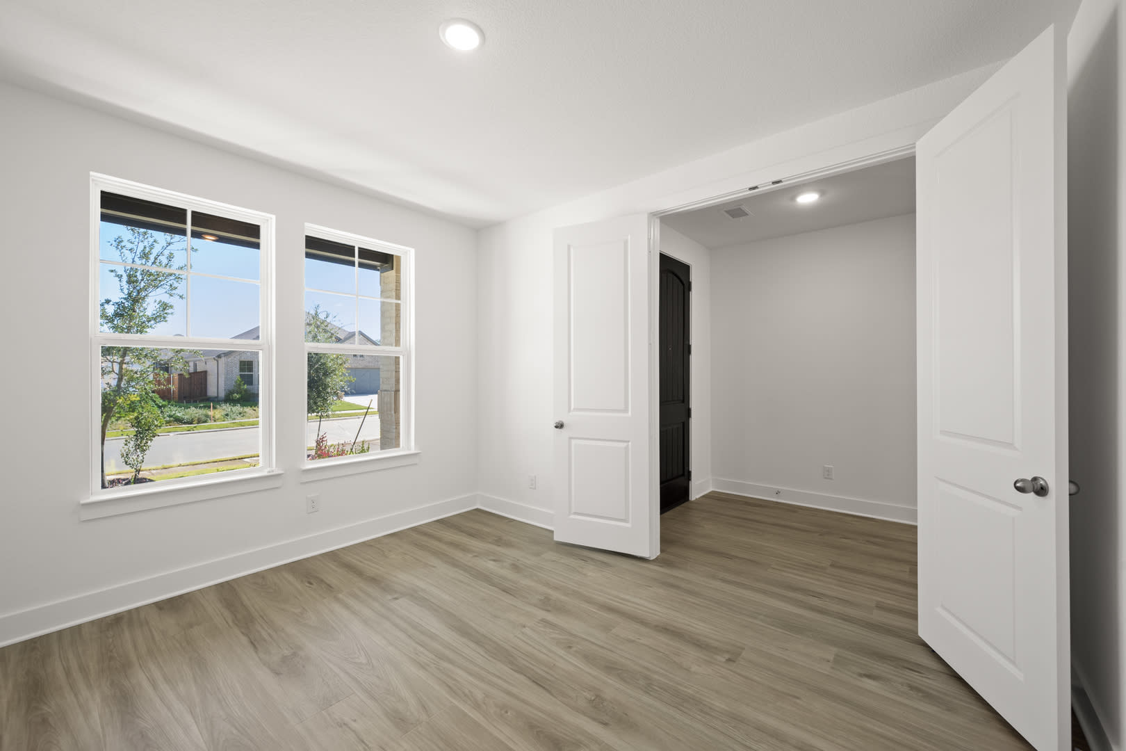 A bright, spacious room with white walls, a large window, and a wooden floor. The room appears to be a bedroom or living space, with a door visible in the background.