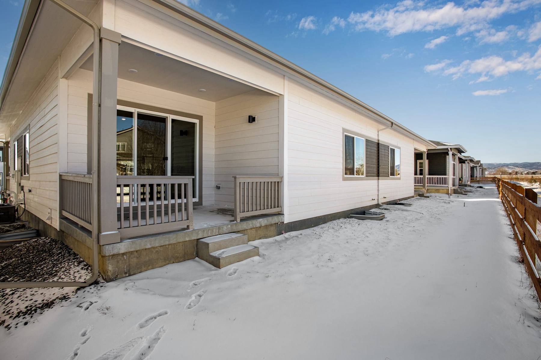 A row of modern, single-story houses with white siding and wooden decks, set against a backdrop of a snowy landscape and a clear blue sky.