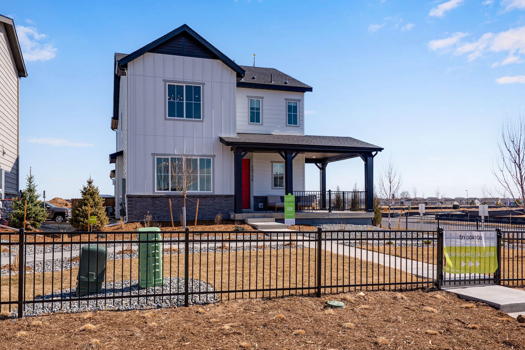A two-story white and gray house with a covered porch, surrounded by a black metal fence and situated in a residential neighborhood with trees and a grassy yard in the foreground.