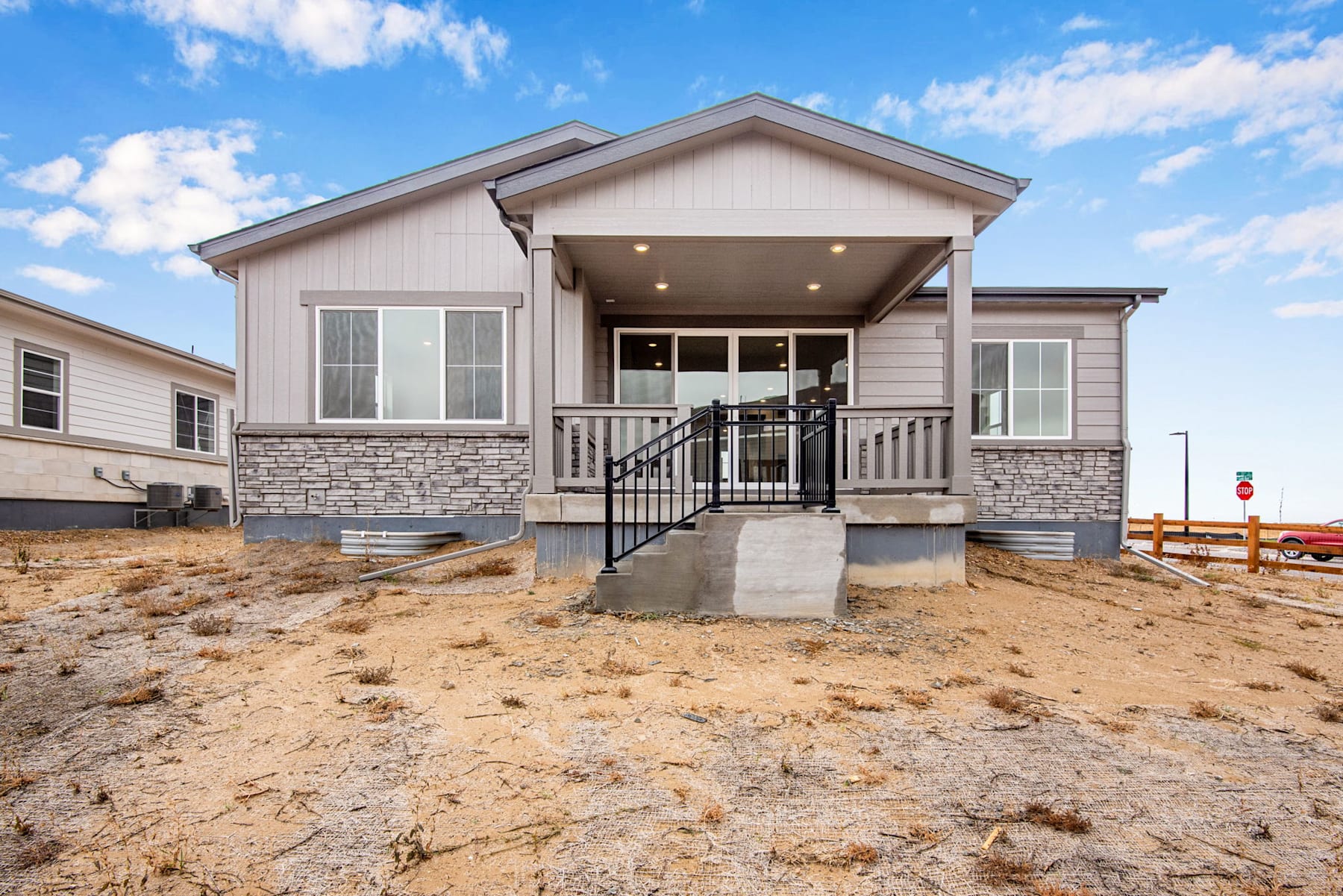A newly constructed single-story house with a covered porch, surrounded by a sandy, barren landscape under a cloudy blue sky.