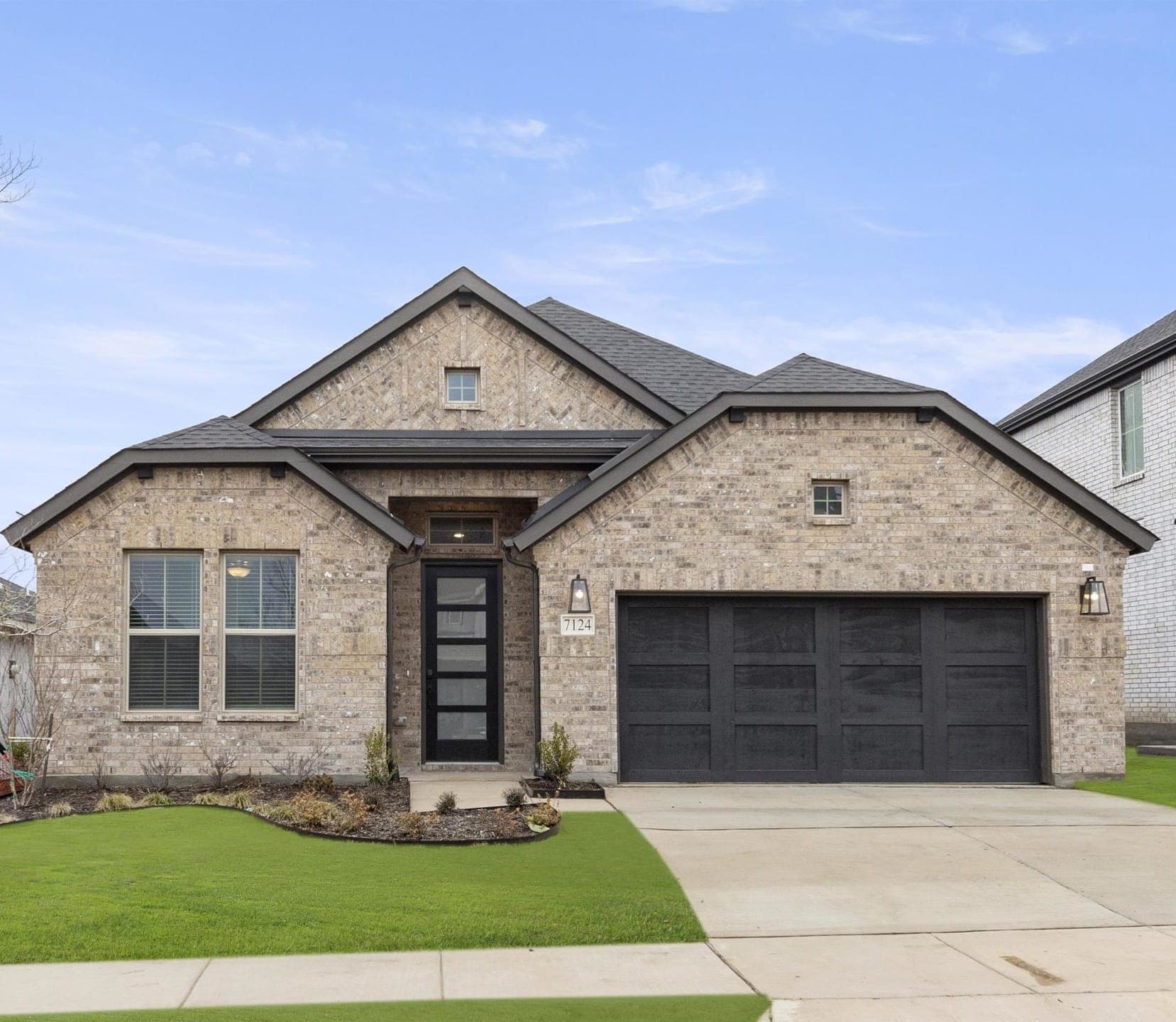 A two-story brick house with a gabled roof, a garage, and a well-manicured lawn in the foreground, set against a clear blue sky.