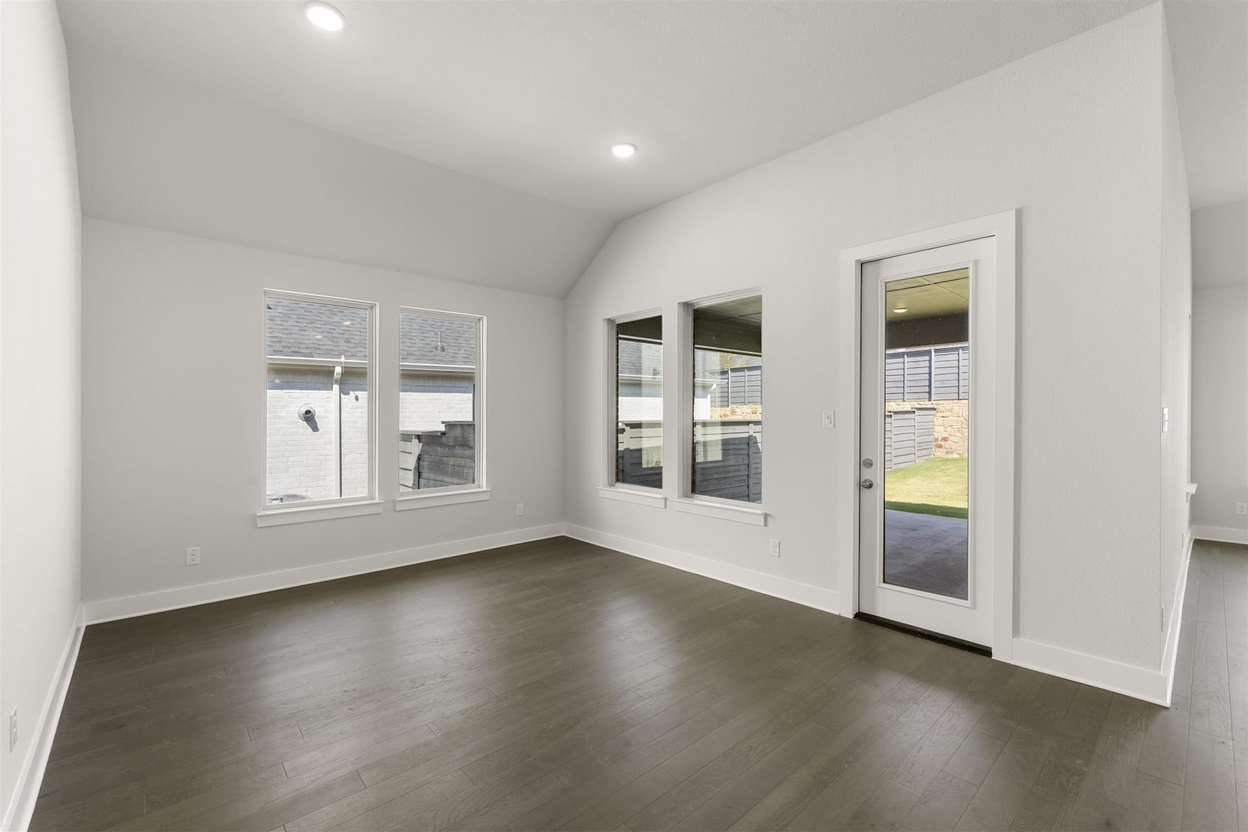 A spacious, well-lit room with white walls, dark hardwood floors, and a glass door leading to an outdoor area.