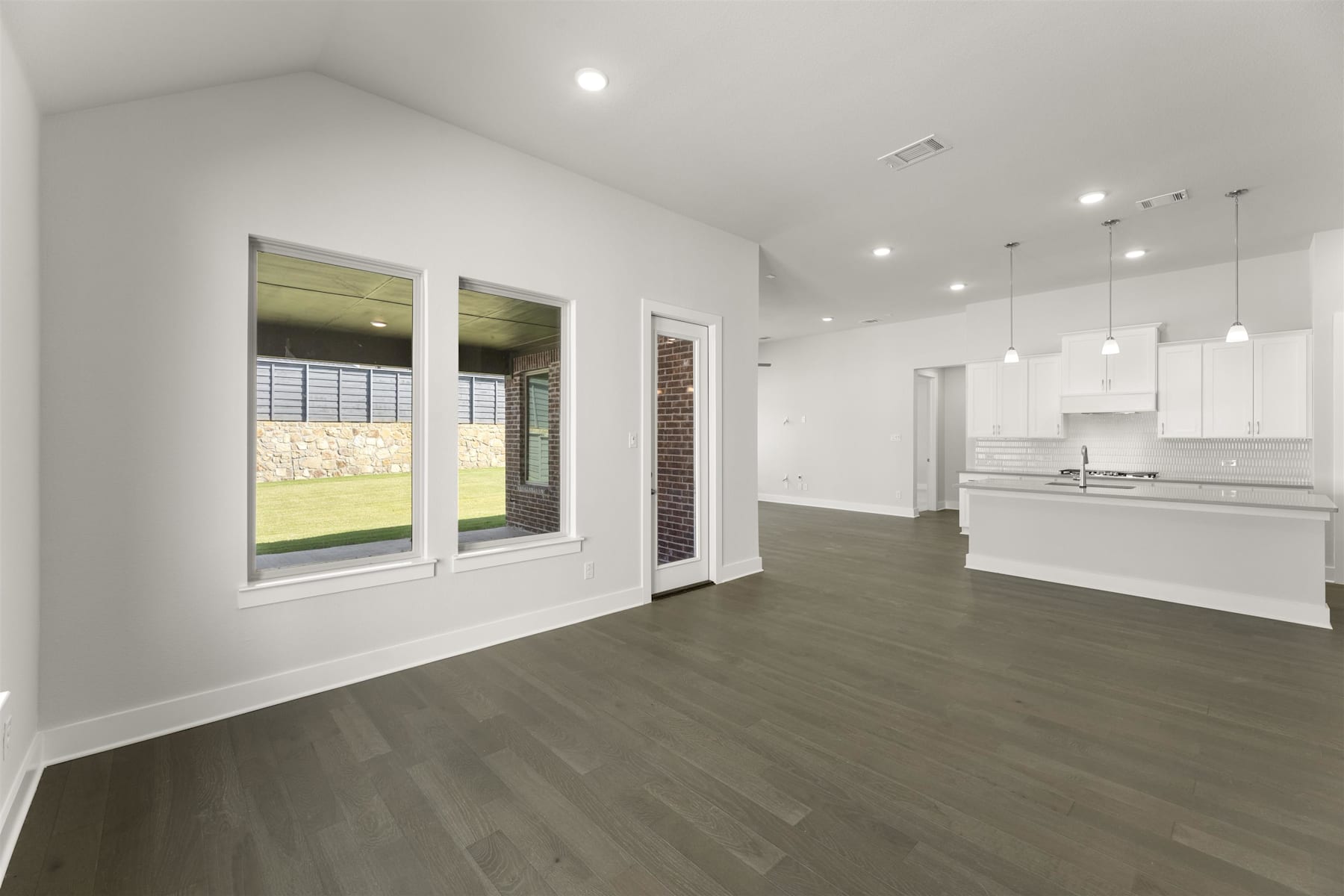 A spacious, modern living room with white walls, dark hardwood floors, and large windows overlooking a grassy outdoor area.